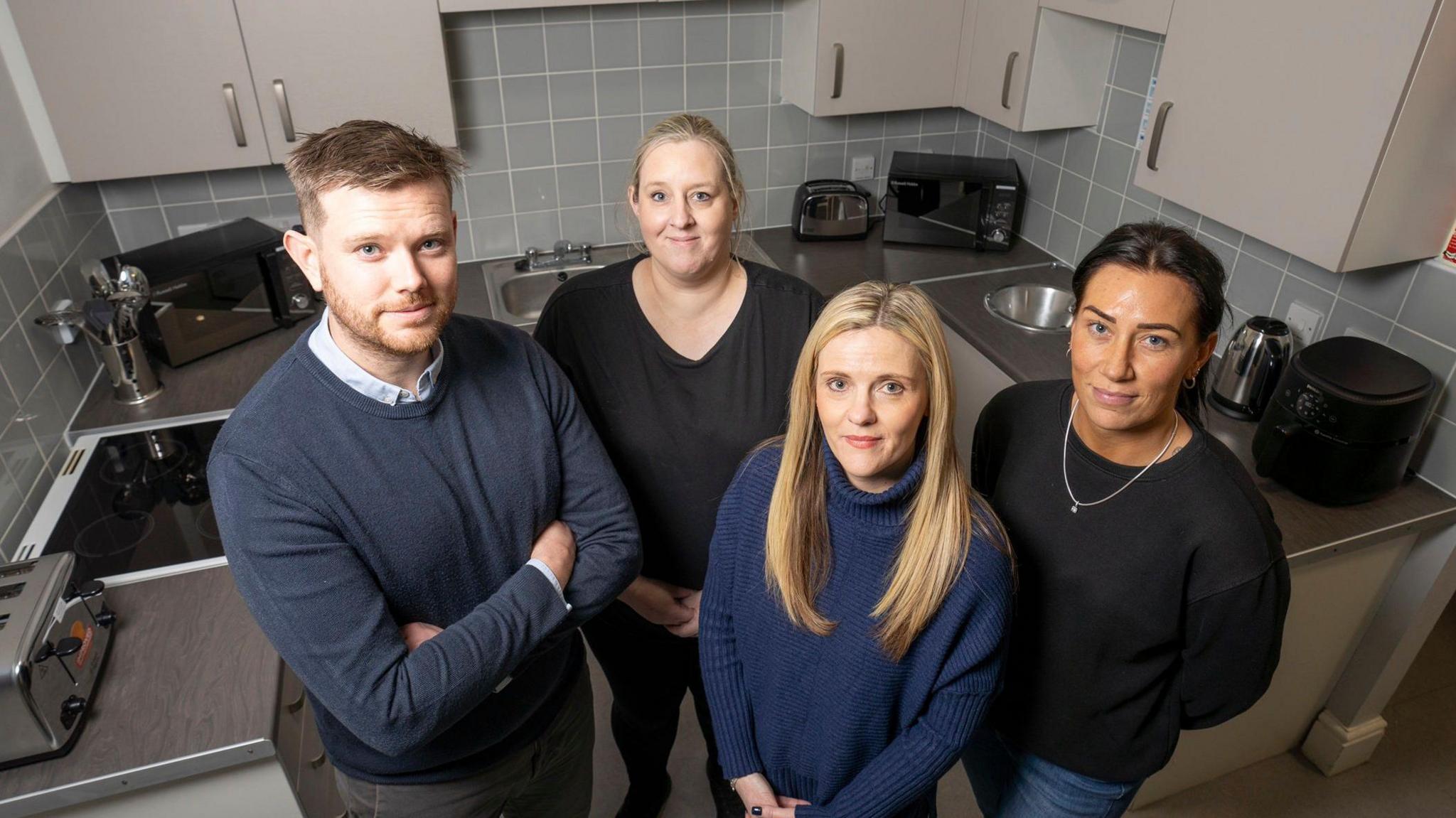 Chris Hepworth, homelessness prevention manager at Durham County Council; Kate Hartley, project lead at Changing Lives; Emma Regan; housing team leader (strategy, partnerships and commissioning) at Durham County Council; and Stacey Lynch, private sector officer at Durham County Council at the kitchen in Teesdale House. Ms Hartley and Ms Lynch are both wearing black jumpers and have their hair in a ponytail. Ms Regan has long blonde hair parted in the middle and is wearing a navy blue jumper. Mr Hepworth has short blonde hair and is wearing a dark blue jumper on top of a shirt. His arms are crossed. Behind the group  there are kitchen benches with two microwave ovens, two toasters and a kettle. There is also a hob and a sink, as well as drawers.