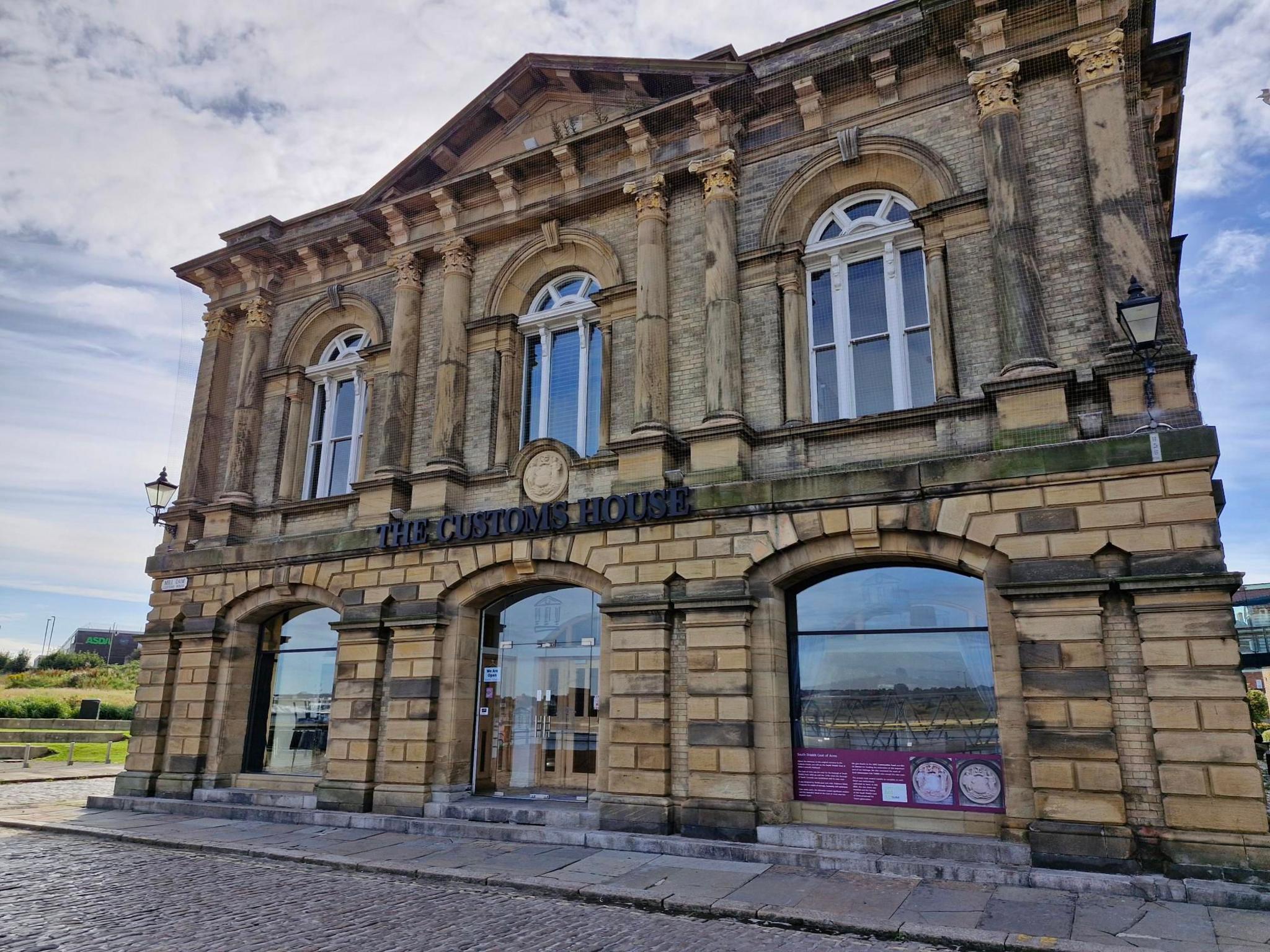 The two-storey building is a grand white stone design with tall arched windows. Front and centre of the building are letter spelling 'The Customs House' with a new white stone crest above it.