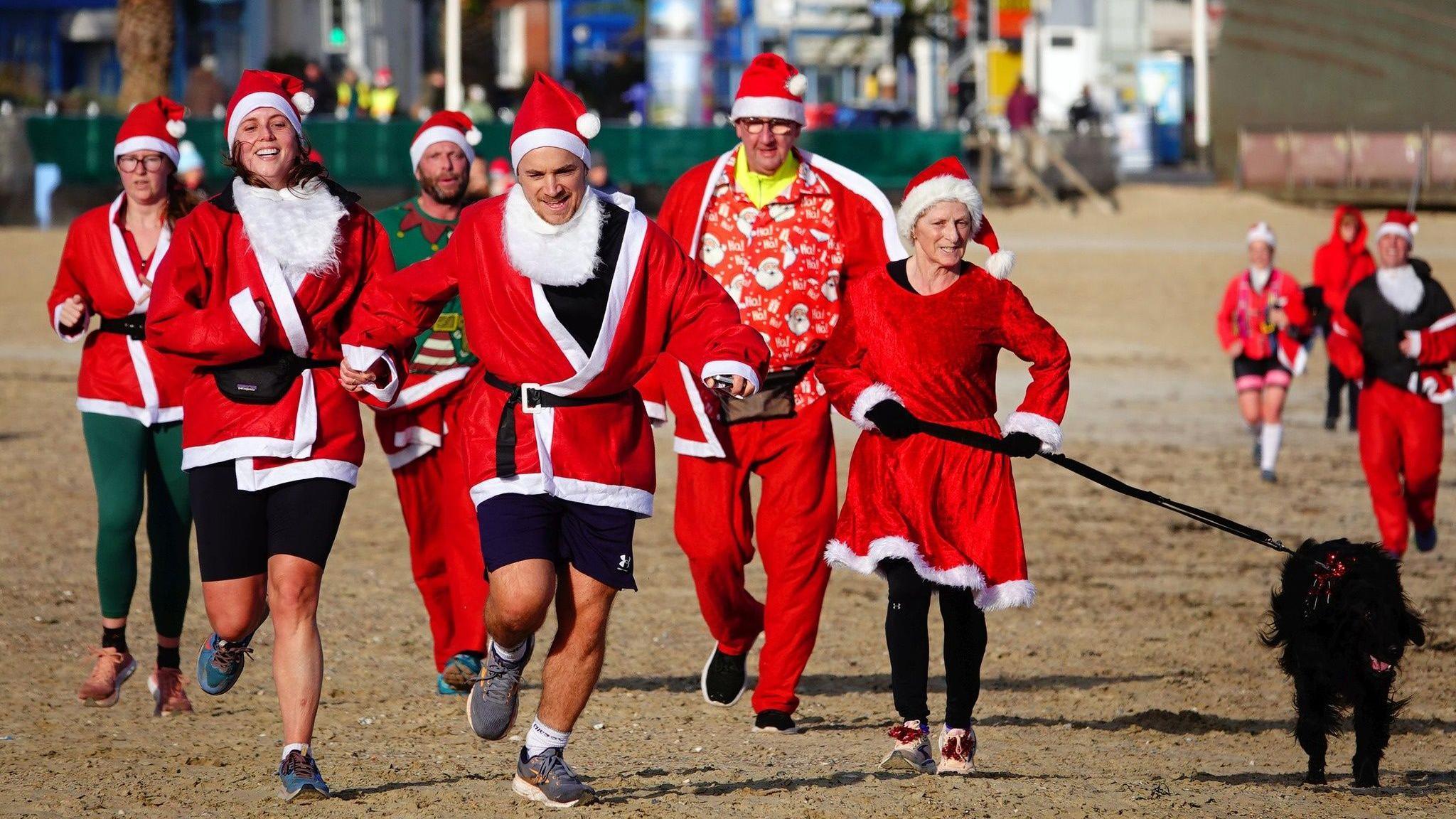 People dressed as santa and a dog running along the beach