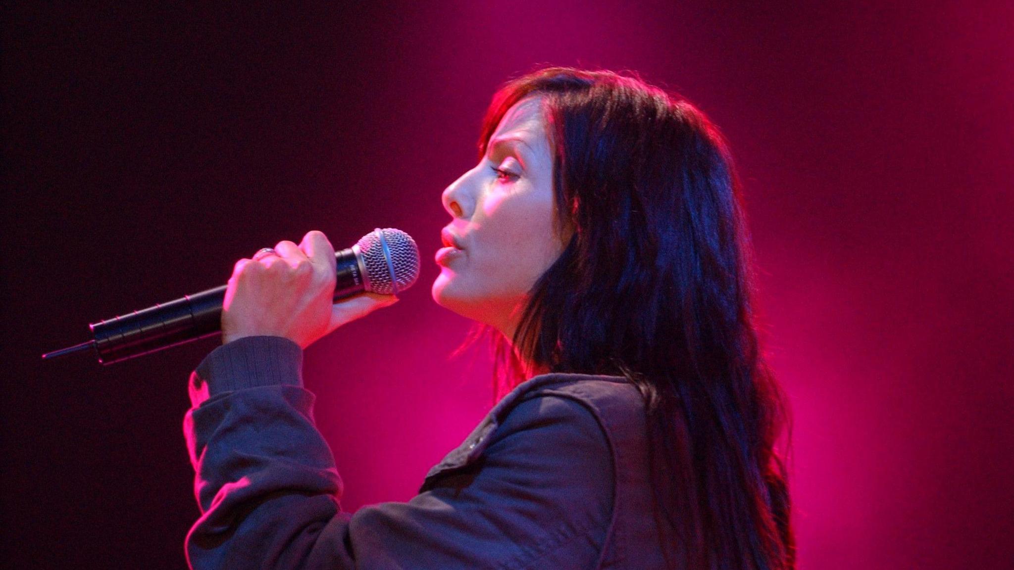 Natalie Imbruglia sings into a microphone. She has long brown hair and a pink light behind her.