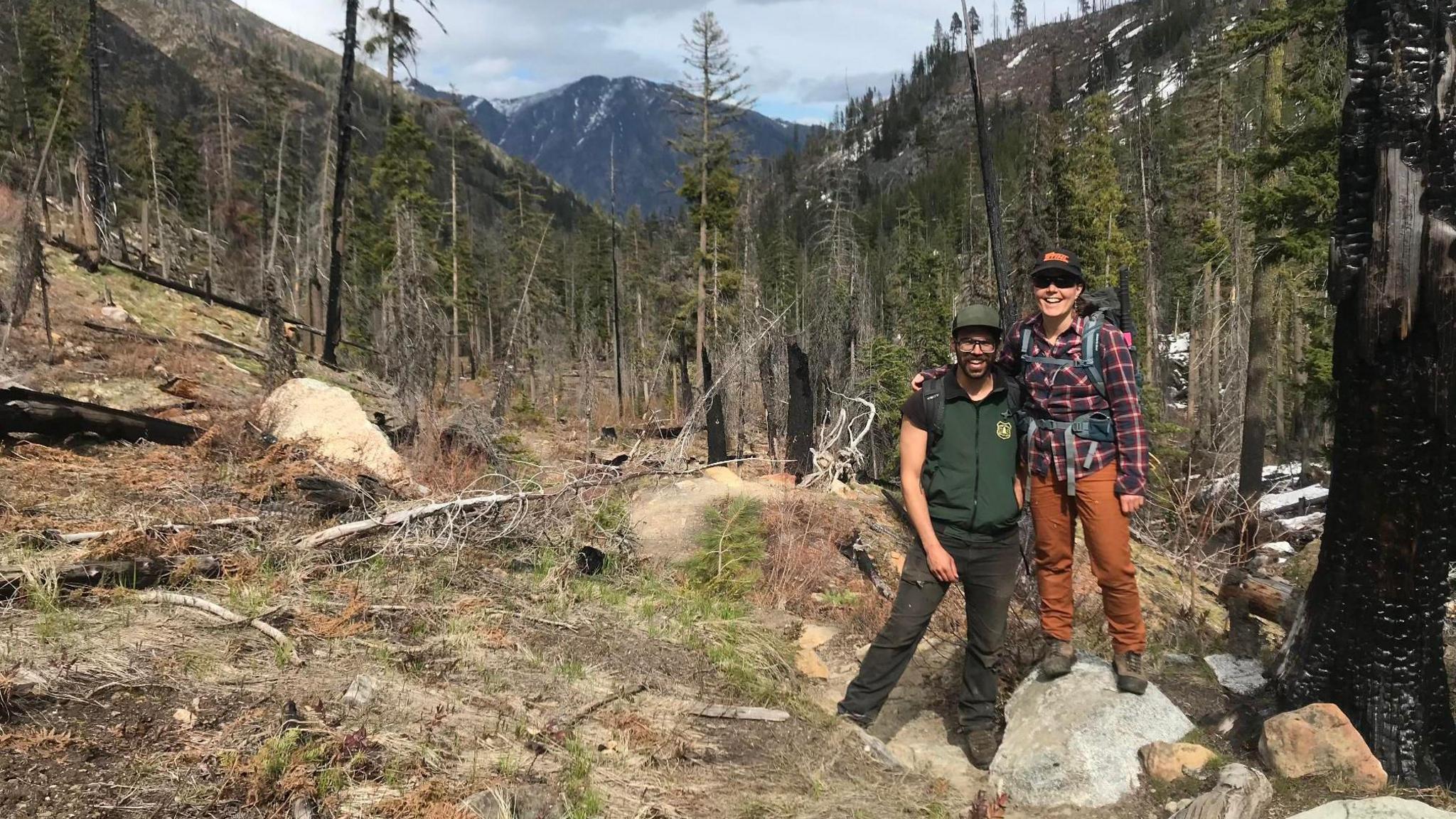 Demetrios and Thompson stand smiling in the middle of a clearing in a hilly forest, with a mountain peak visible in the background. Demetrios has a beard and is wearing a green sport vest and brown work pants and brown hiking boots, with a baseball cap shielding his eyes. Thompson is standing next to him on a rock so that she is taller, wearing orange work pants, a red flannel shirt, a baseball hat and a hiking backpack. 