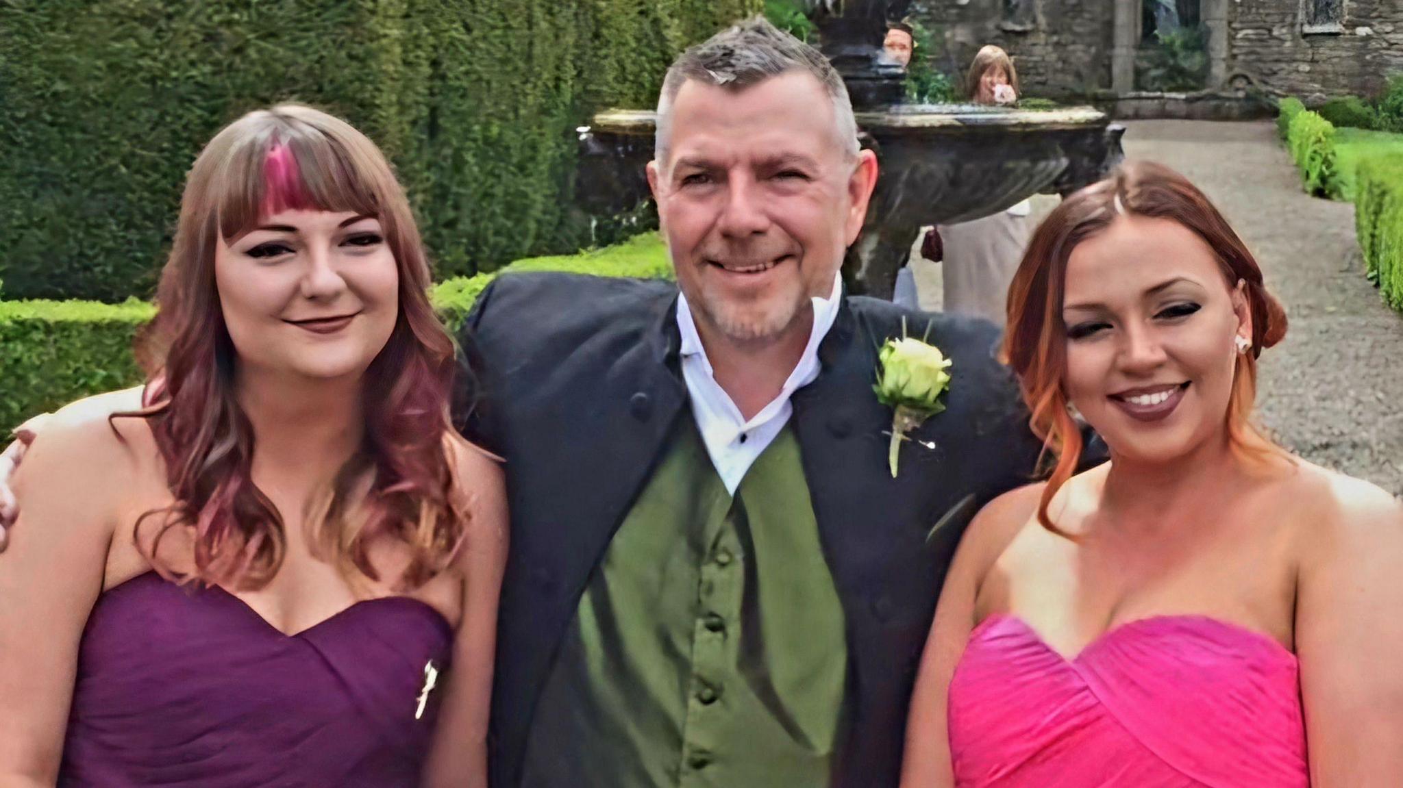 Brian Place wearing a suit and green waistcoat standing with his arms around his two daughters. The woman on the left has long, curly, brown and pink hair and is wearing a purple dress. The woman on the right has brown and orange curly hair and is wearing a pink dress. 