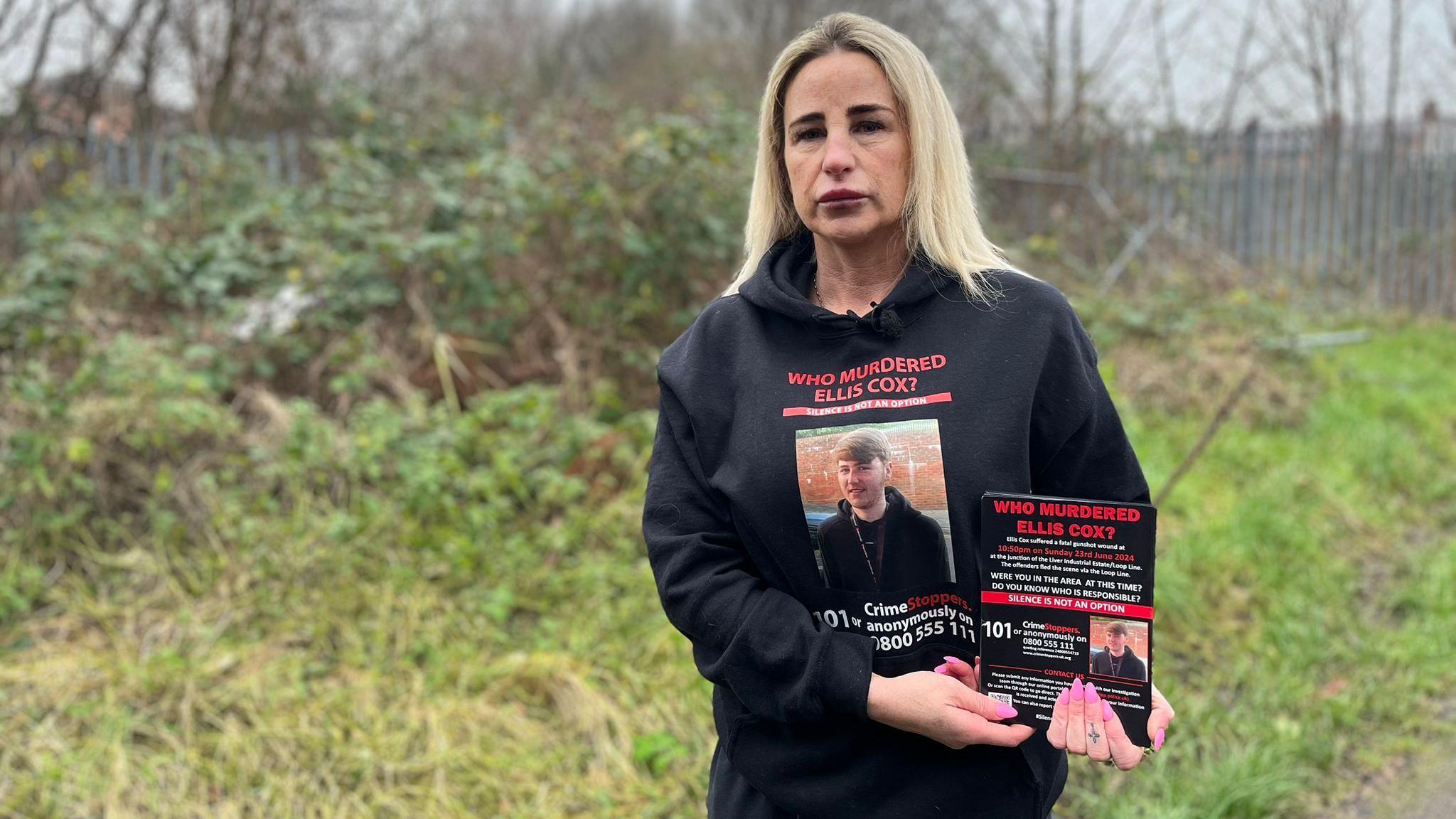 Carolyn Cox stands on a path next to a grassed area. She is holding flyers appealing for help in finding her son's killer. She has long blonde hair and is wearing a black sweatshirt which includes the words 'who murdered Ellis Cox?' and a photo of her son.