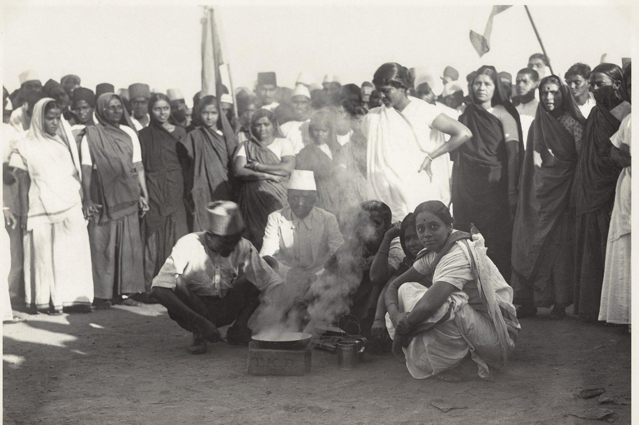 Three women and two men are seen crouched on the shores of Mumbai's Chowpatty Beach, ready to make contraband salt. A crowd, mostly comprising women, stands behind them and watches on.