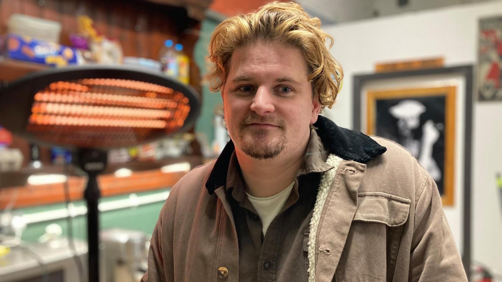 A young man with curly fair hair smiles shyly. He wears a goatee beard and a thick winter coat