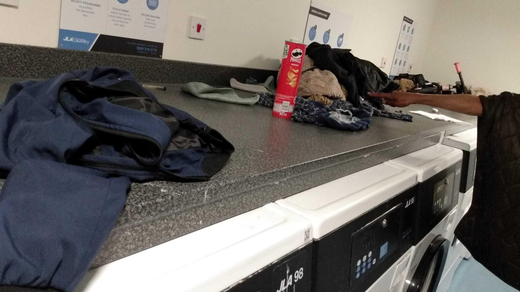 A grey marble effect kitchen worktop is covered in rubbish, empty Pringles crisps packets and clothes.