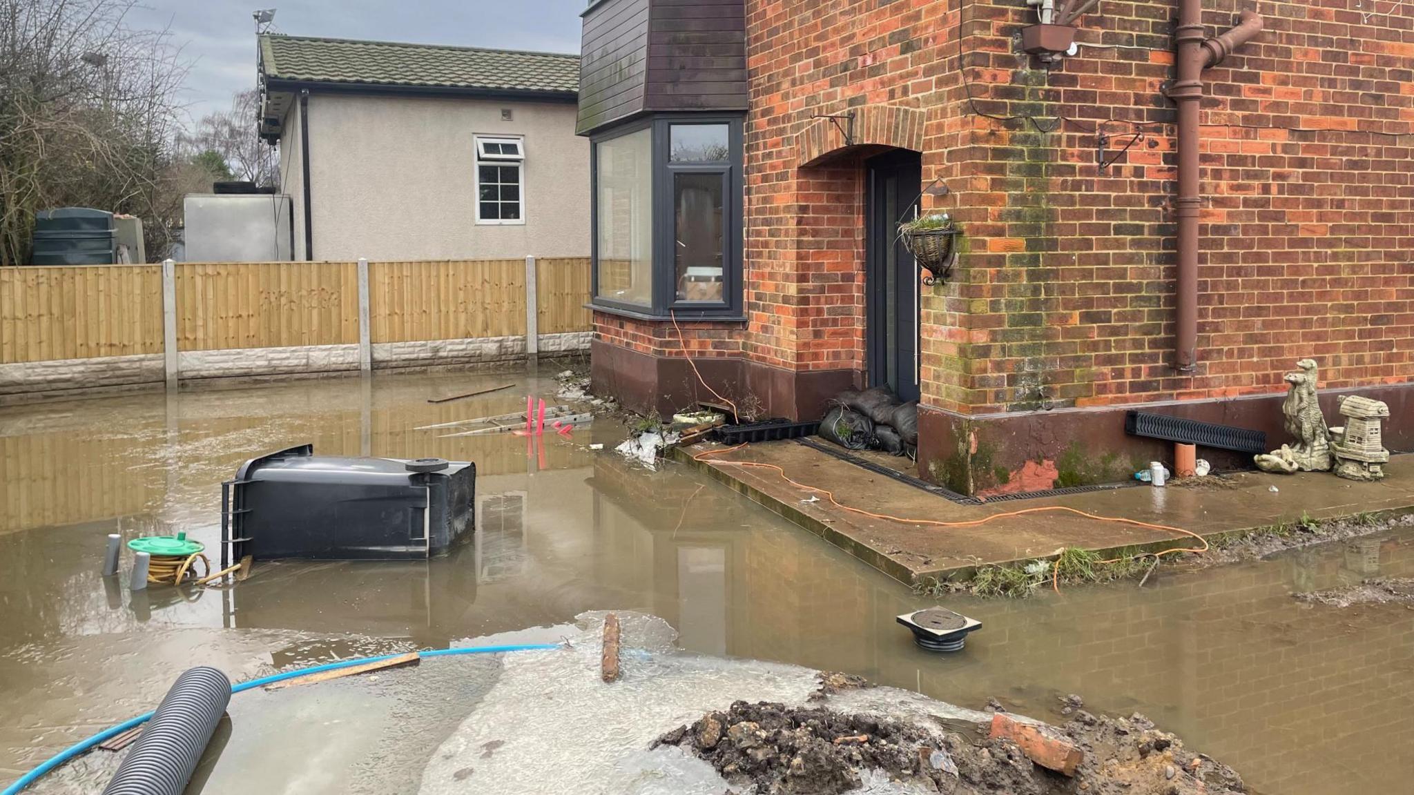 Floodwater surrounds a house with sandbags piled up at the door. A wheelie bin is tipped over on its side in the garden.