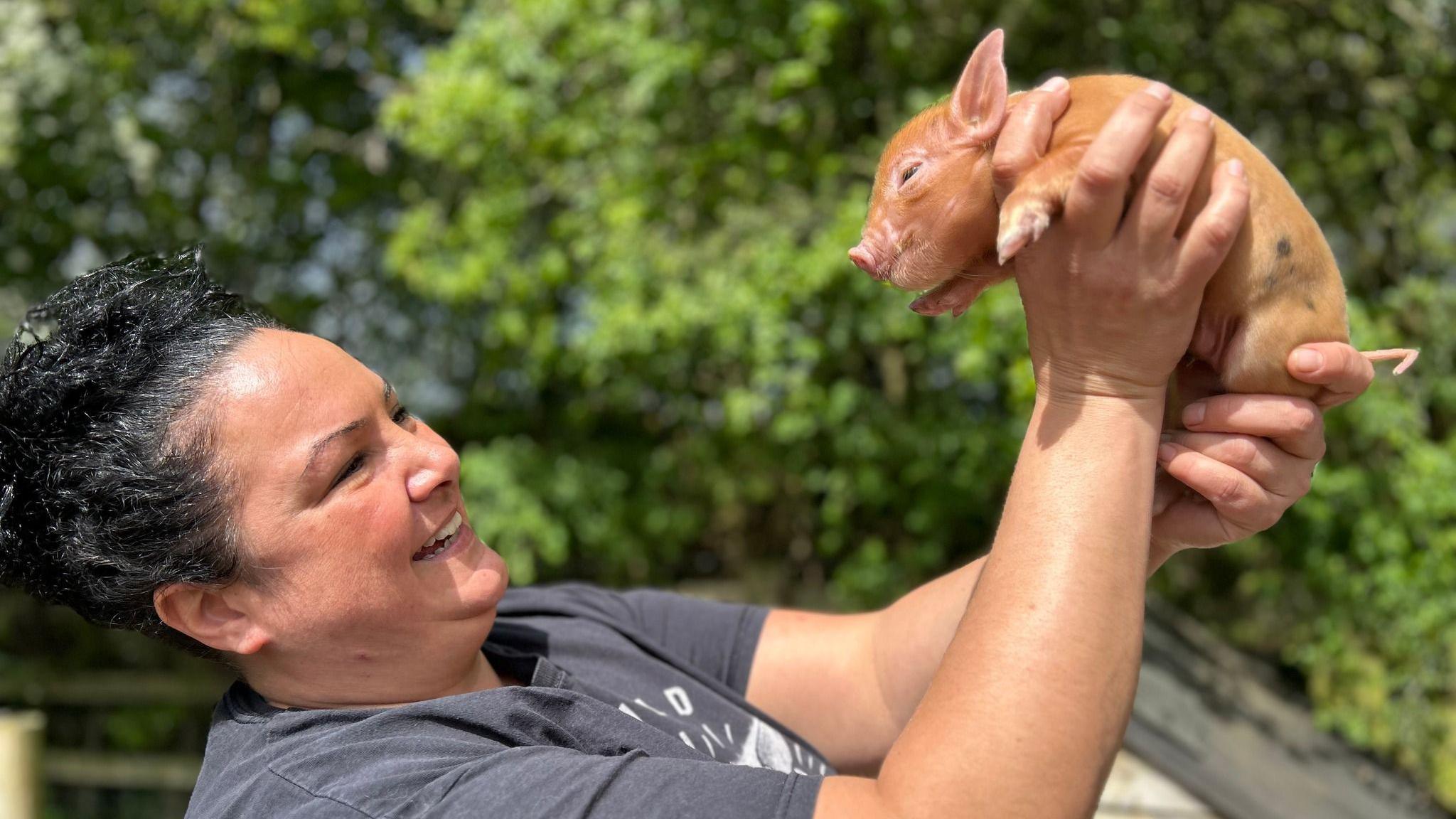 A woman holding a micro pig in the air wearing a grey top 