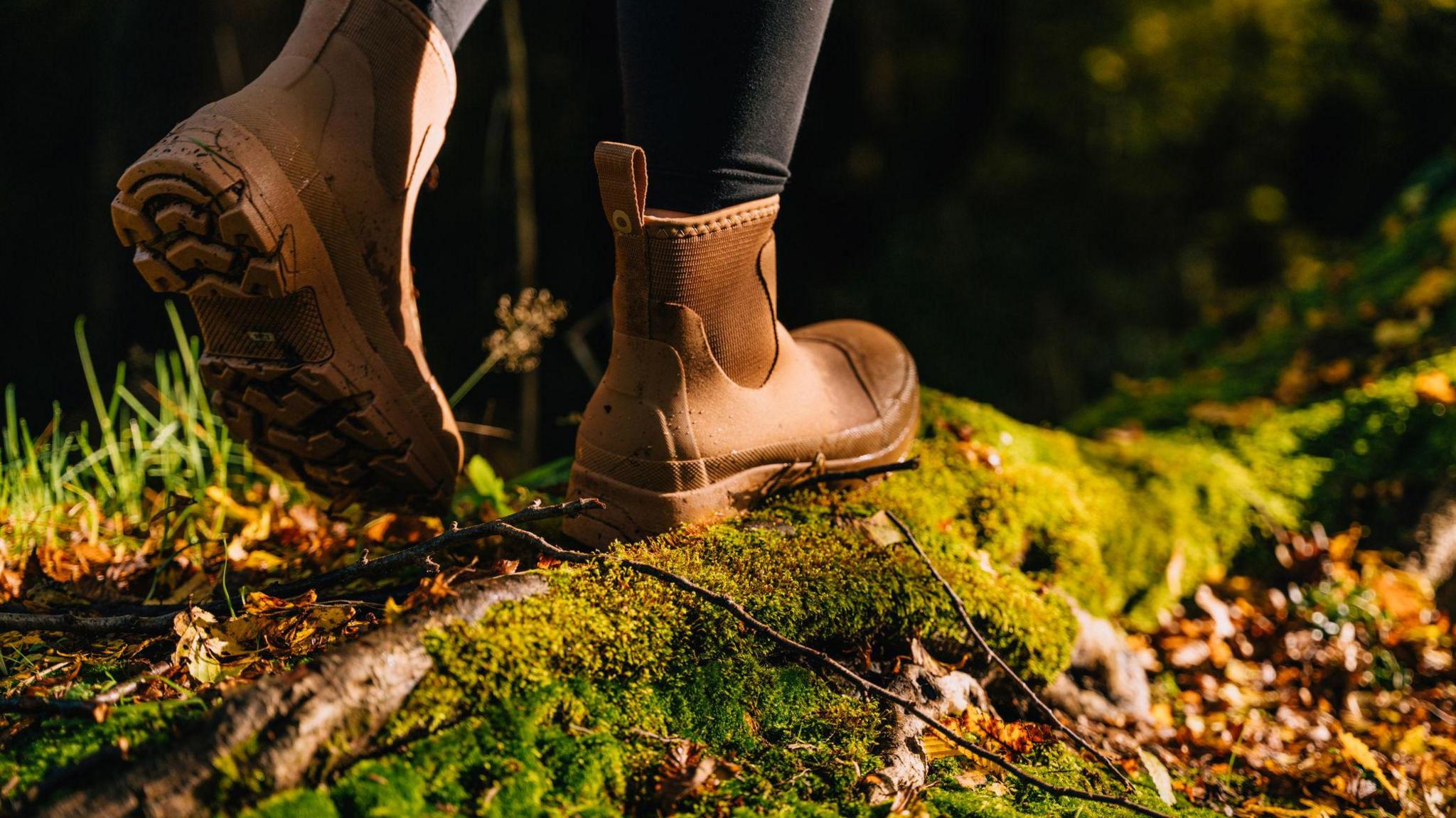 A pair of feet in brown ankle-length boots walk along what appears to be moss-covered log. Blades of grass can also be seen. The person's legs can be seen in black leggings.