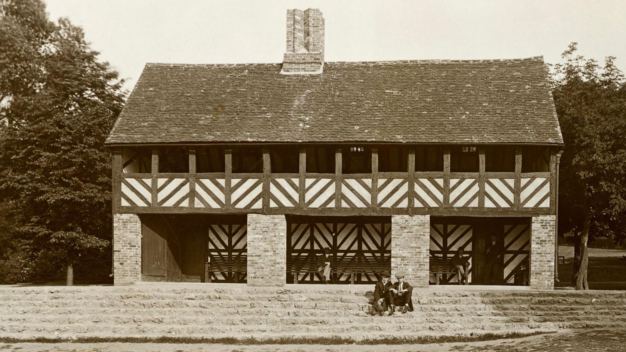 An old black and white photo of a timber-framed pub building situated in a park with steps leading up to it. Two people wearing hats and suits are sitting together on one of the steps.