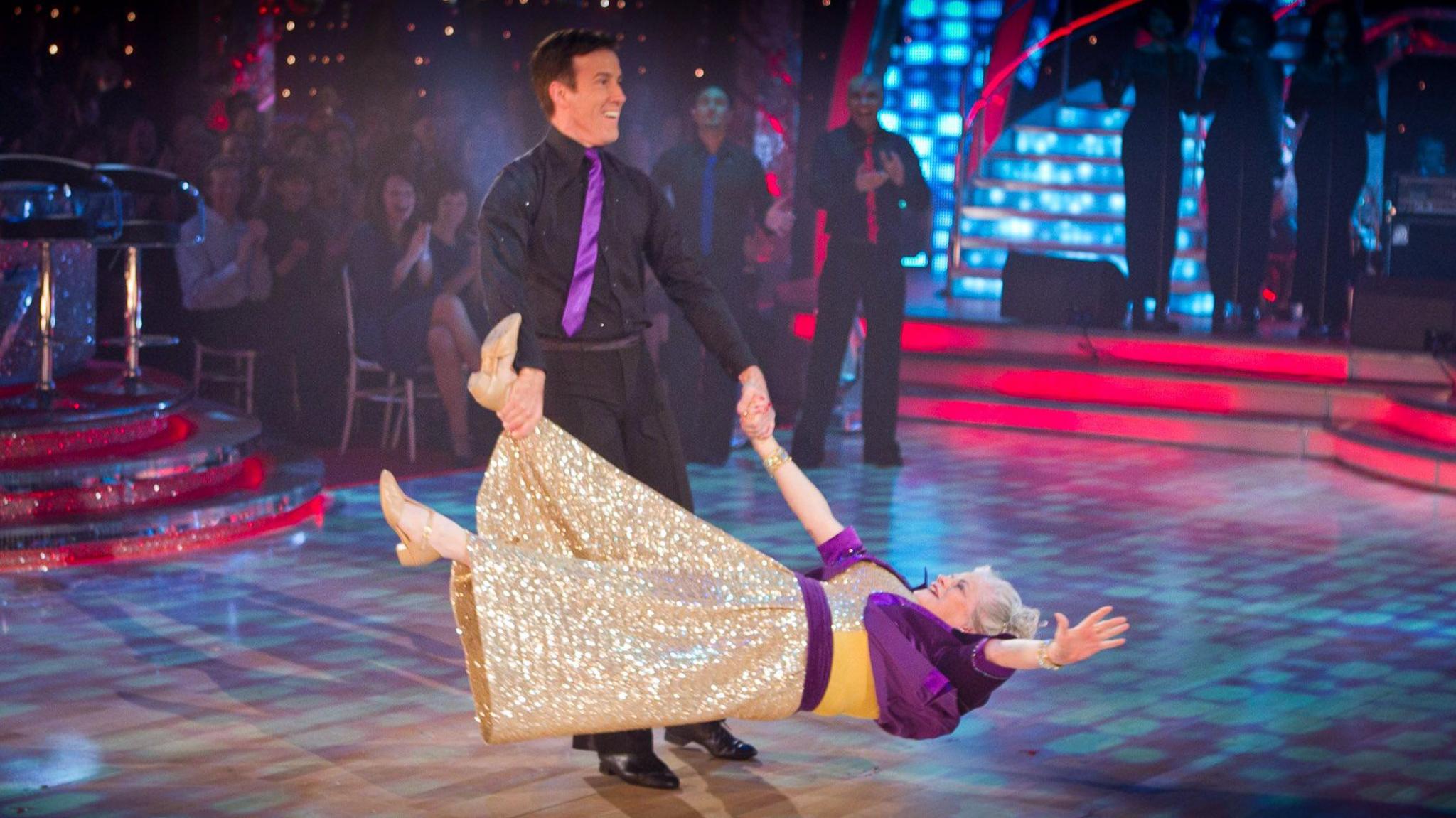 A picture of Widdecombe and Du Beke dancing in the Strictly Come Dancing studio. Anton Du Beke wearing a black shirt and trousers and a purple tie. He is spinning Ann Widdecombe around, holding her by the hand and foot. Widdecombe is wearing a glittery golden dress with purple sleeves.