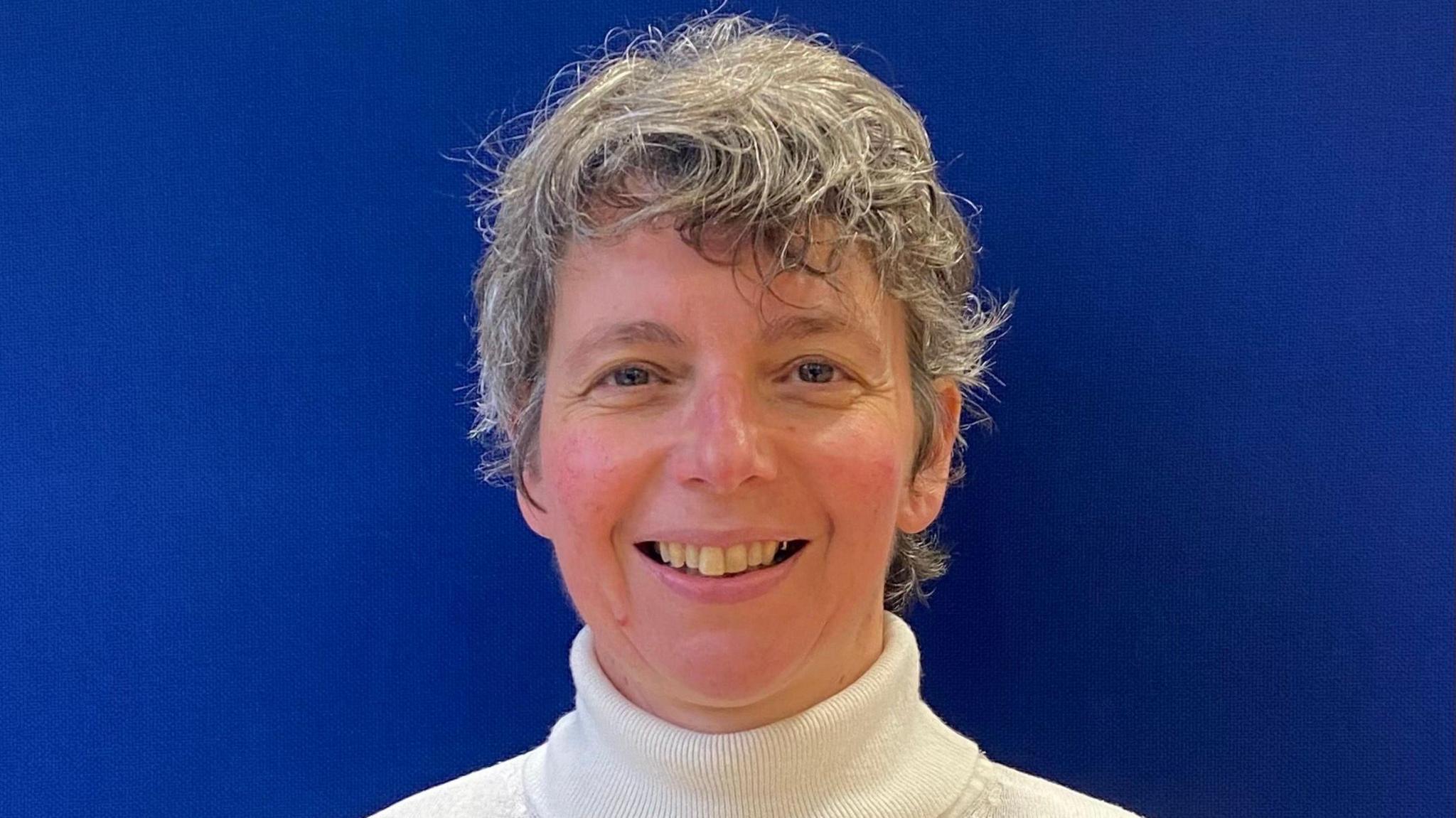 Kirstie Cook standing in front of a blue wall. She is wearing a white turtle necked jumper and is looking straight at the camera and smiling. She has short fair hair. 