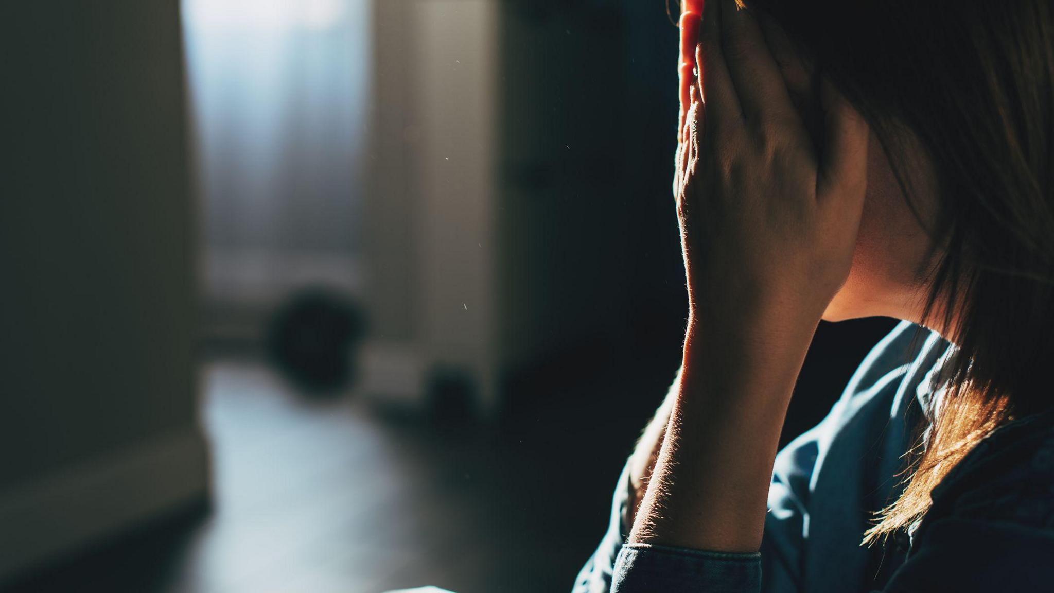 A distressed woman sitting on the floor of a dark room with her head in her hands.  She has dark shoulder-length hair and is wearing a blue top.  There is a window in the background.