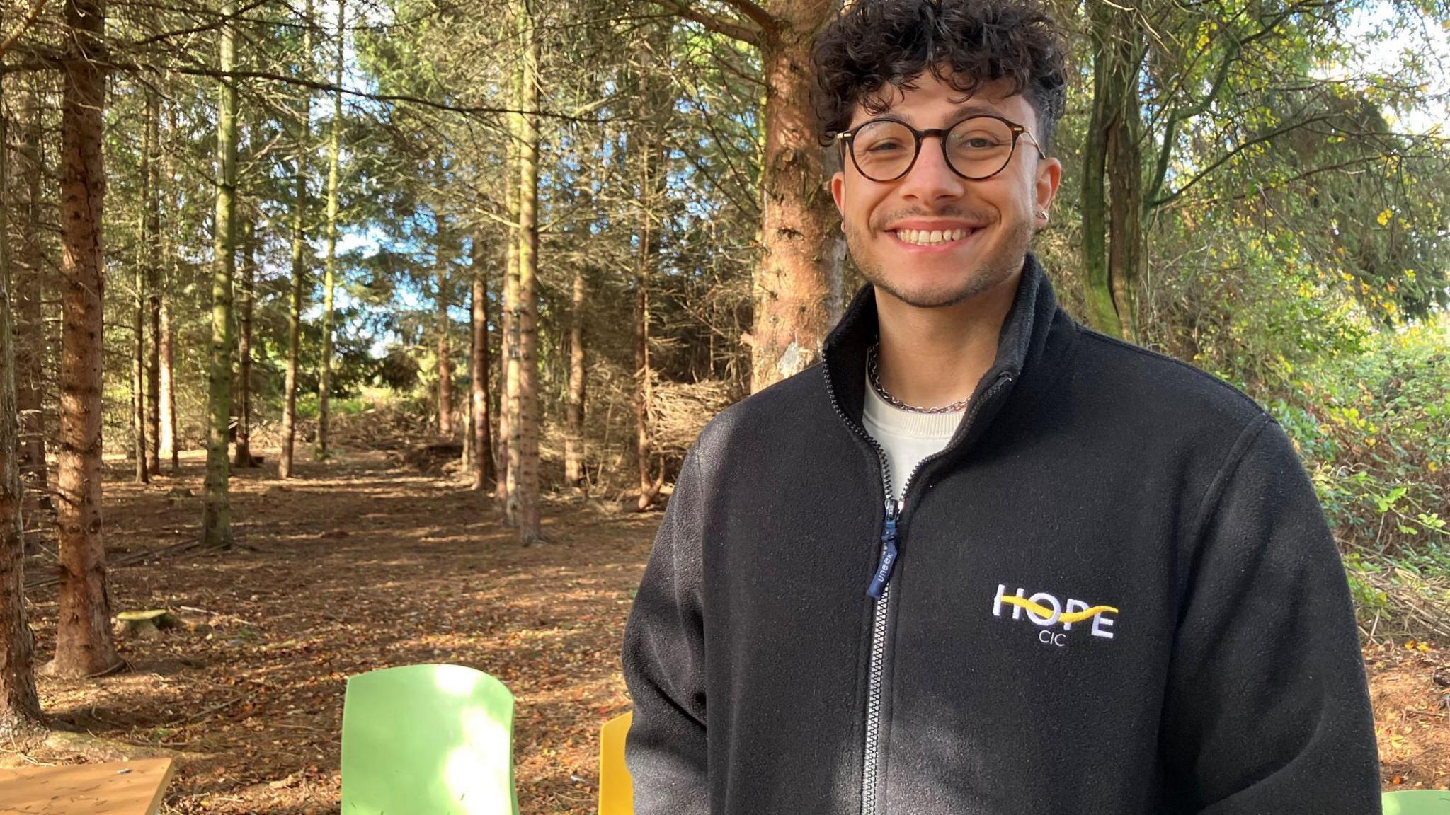 Tomas Mountain wears spectacles and a black fleece. He is stood in a clearing surrounded by trees. There are golden leaves on the ground. Chairs and a table are in view.