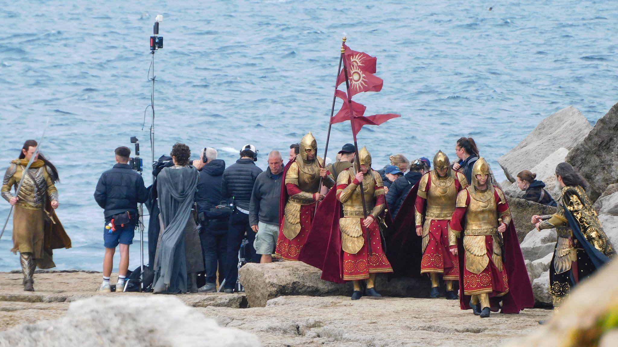 Actor Benjamin Walker as Gil-galad and other actors portraying soldiers filming a scene of The Rings of Power around filming crews by the sea. It is overcast.