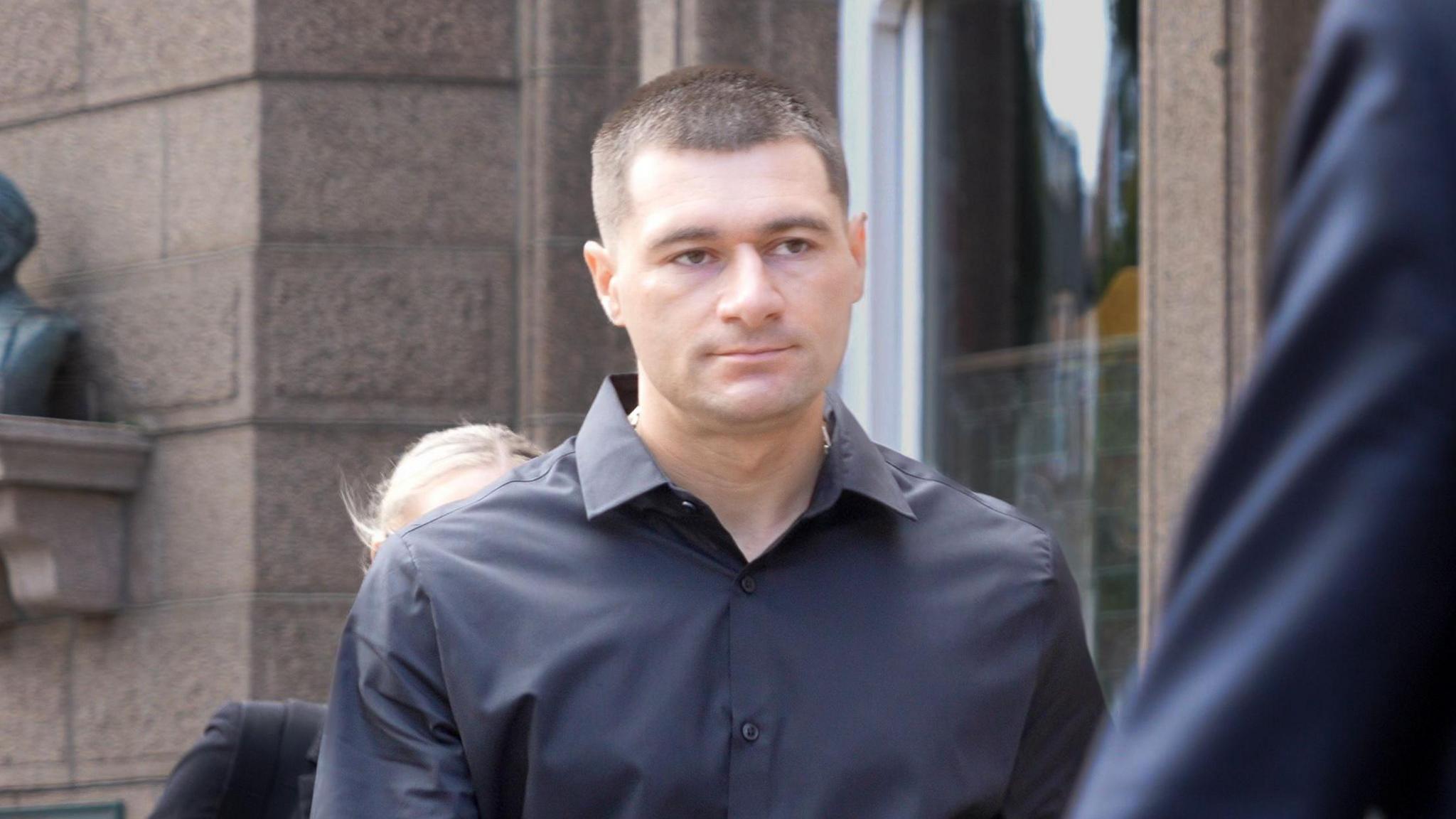 Artur Sevash-Zade wearing a black shirt walks into court. He has short black hair and is clean shaven.
