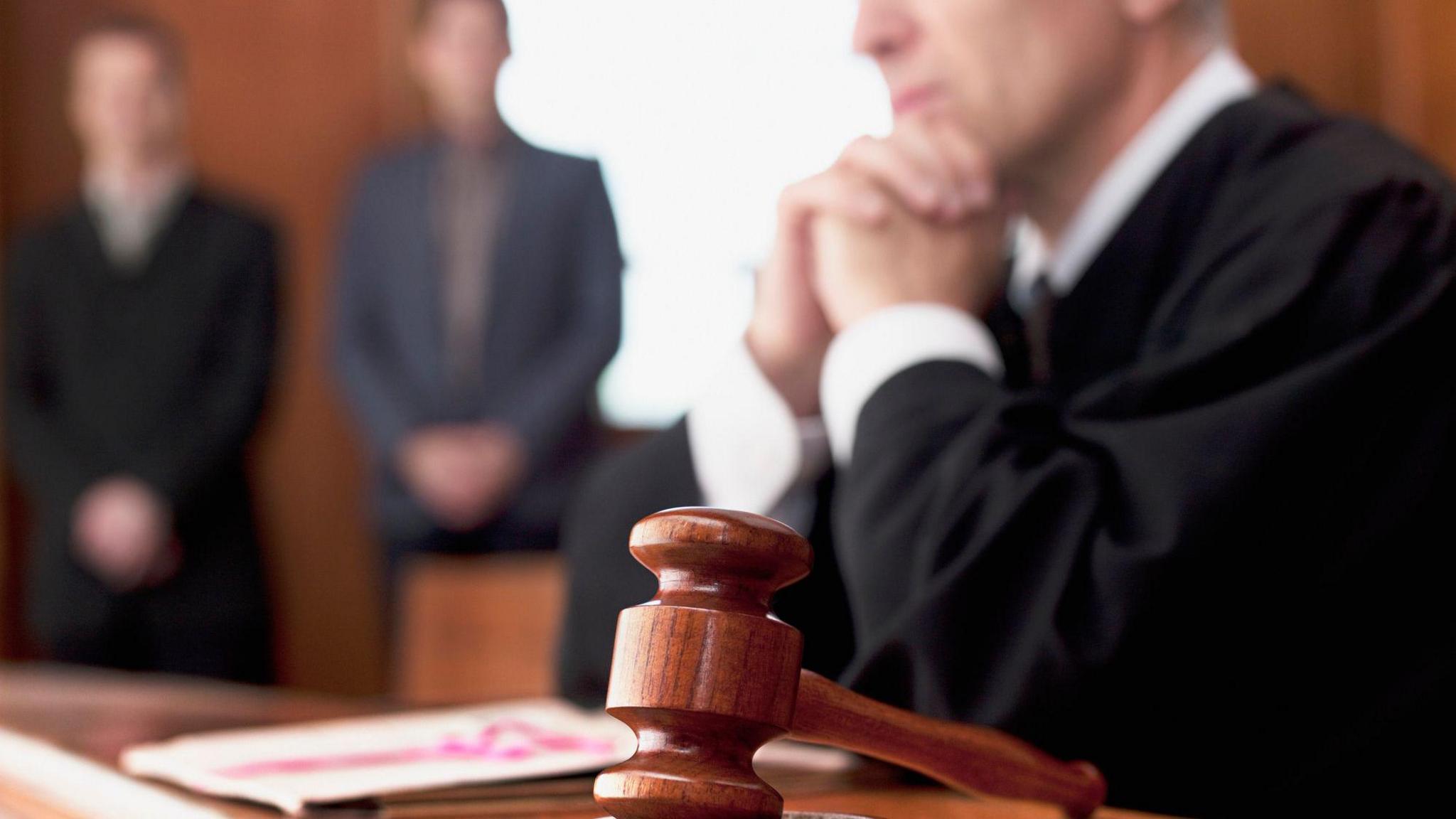 Stock photo of a judge and gavel in court. There's a shot of a slightly out of focus man dressed in black as a judge. There's two people standing in the background in suits.