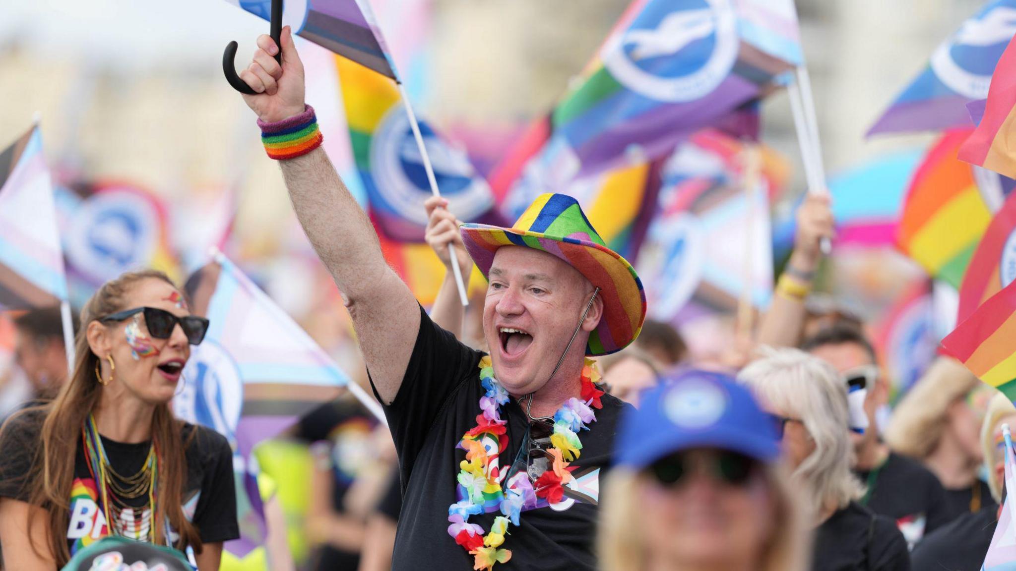 A man with a rainbow hat on. He is smiling towards a crowd of people.