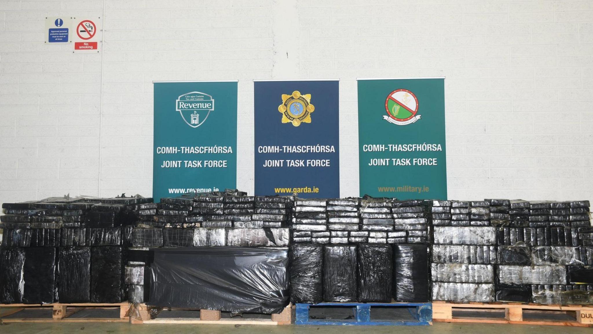 Four pallets have been lined up in a large warehouse. Each is packed with blocks of plastic wrapped packages shaped like bricks. Behind the drug haul are three signs each reading 'joint task force' in Irish and English. It also has a website address for Gardai