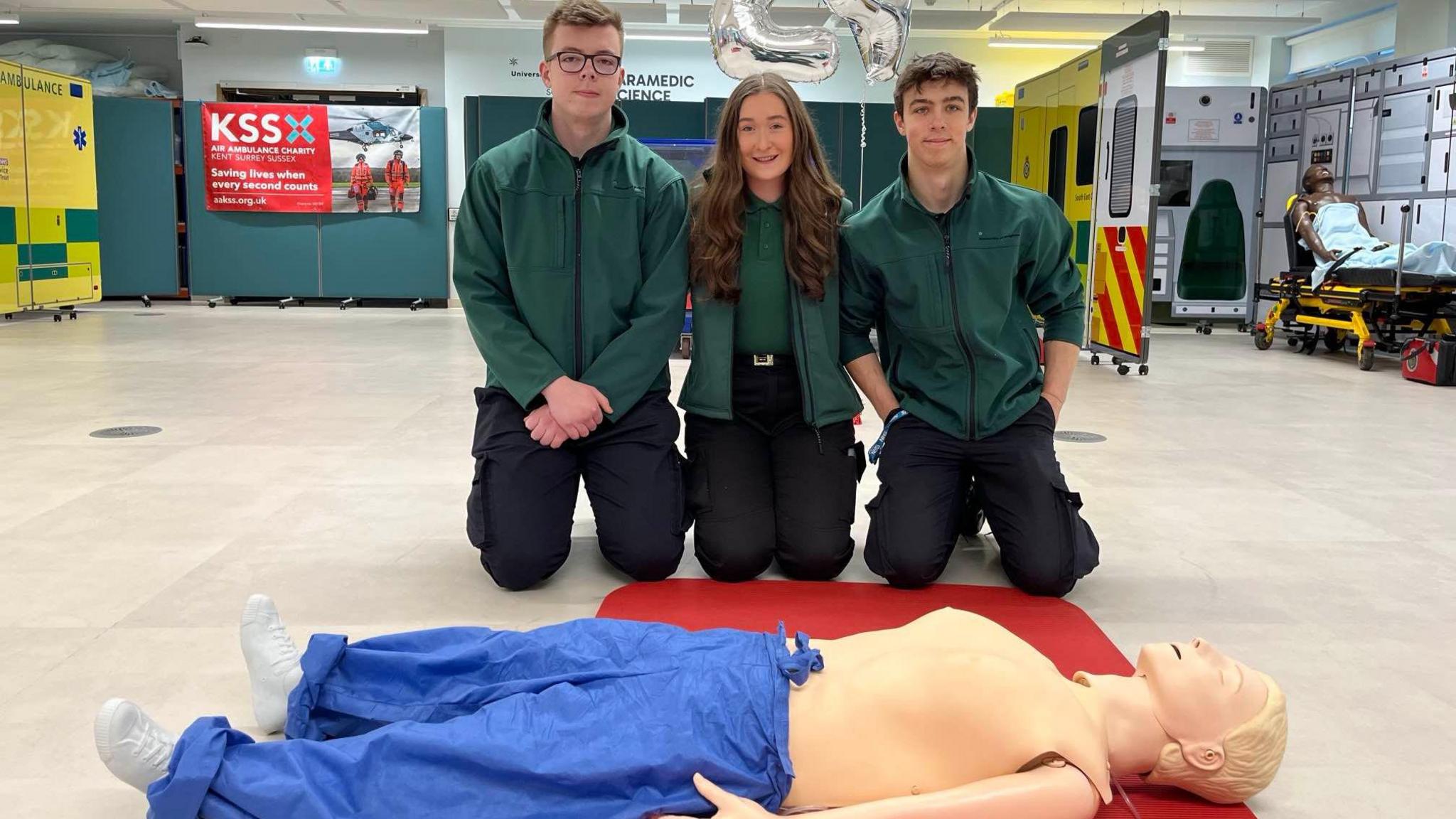 two young men and a young woman all wearing dark green jackets and black trousers kneel in front of a CPR dummy