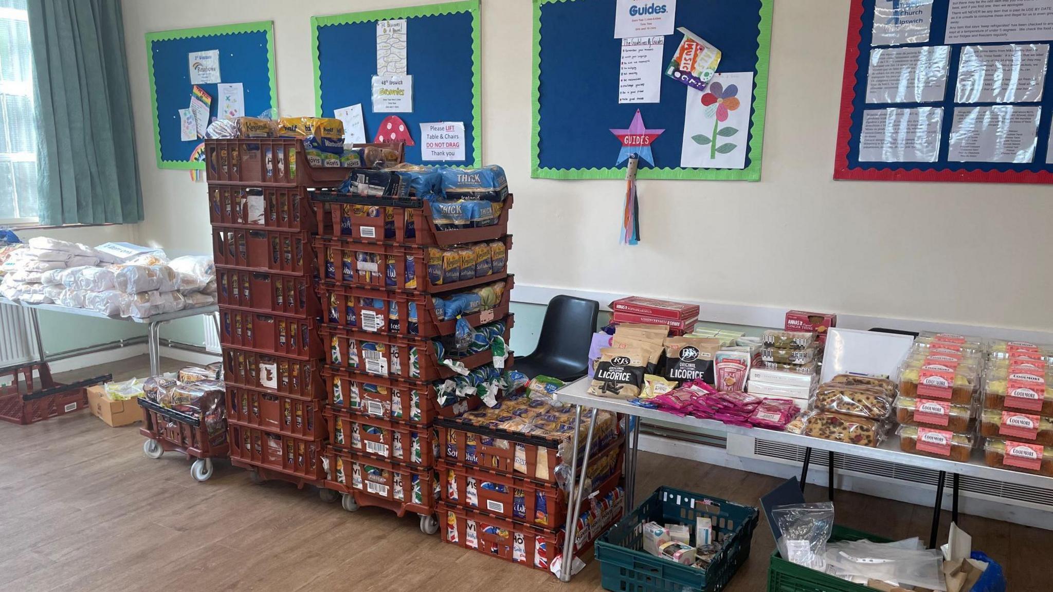 A room with stacked crates and two tables stocked with food items, such as bread, licorice and cakes. 