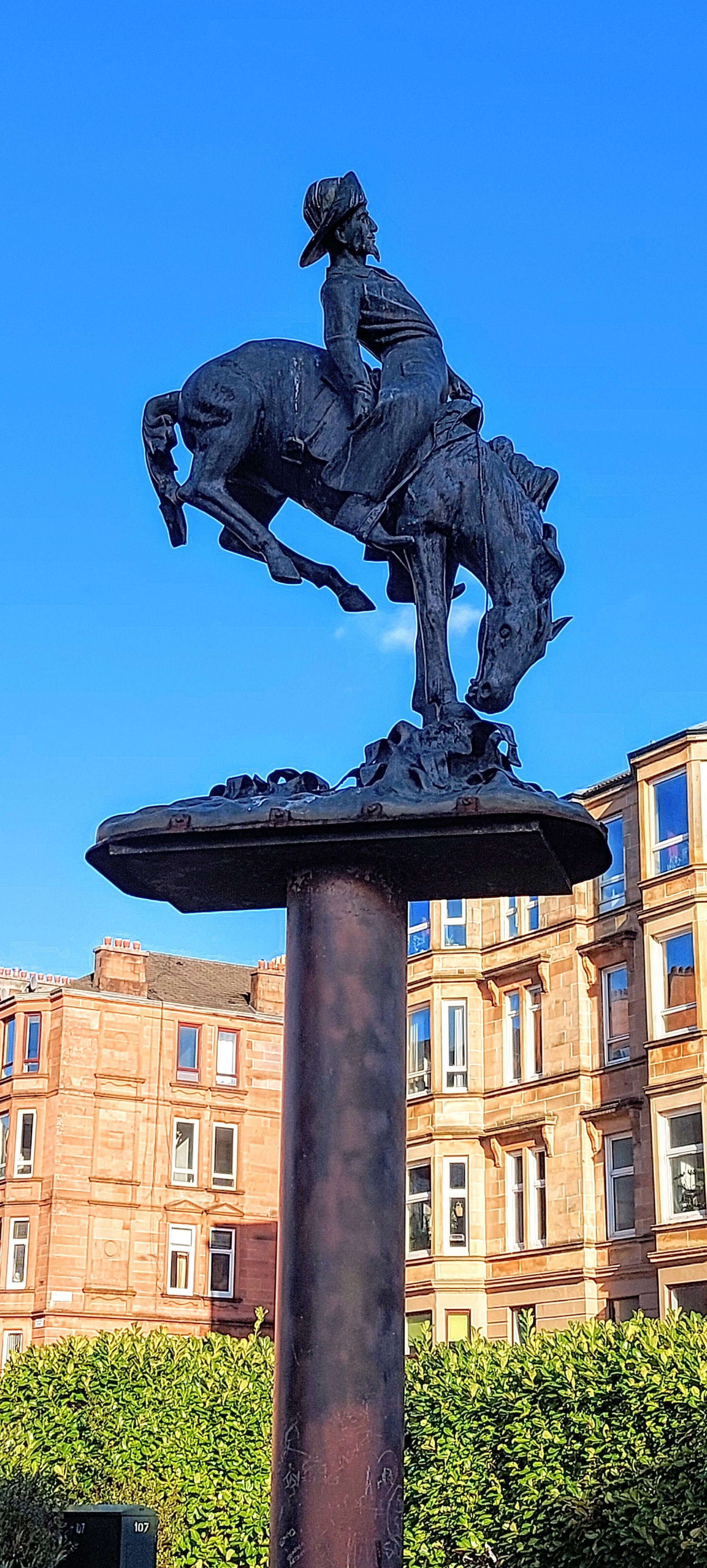 A statue of Buffalo Bill trying to stay on a bucking horse stands on a pedestal in front of some old stone tenement style buildings and a hedge under a blue, blue sky.