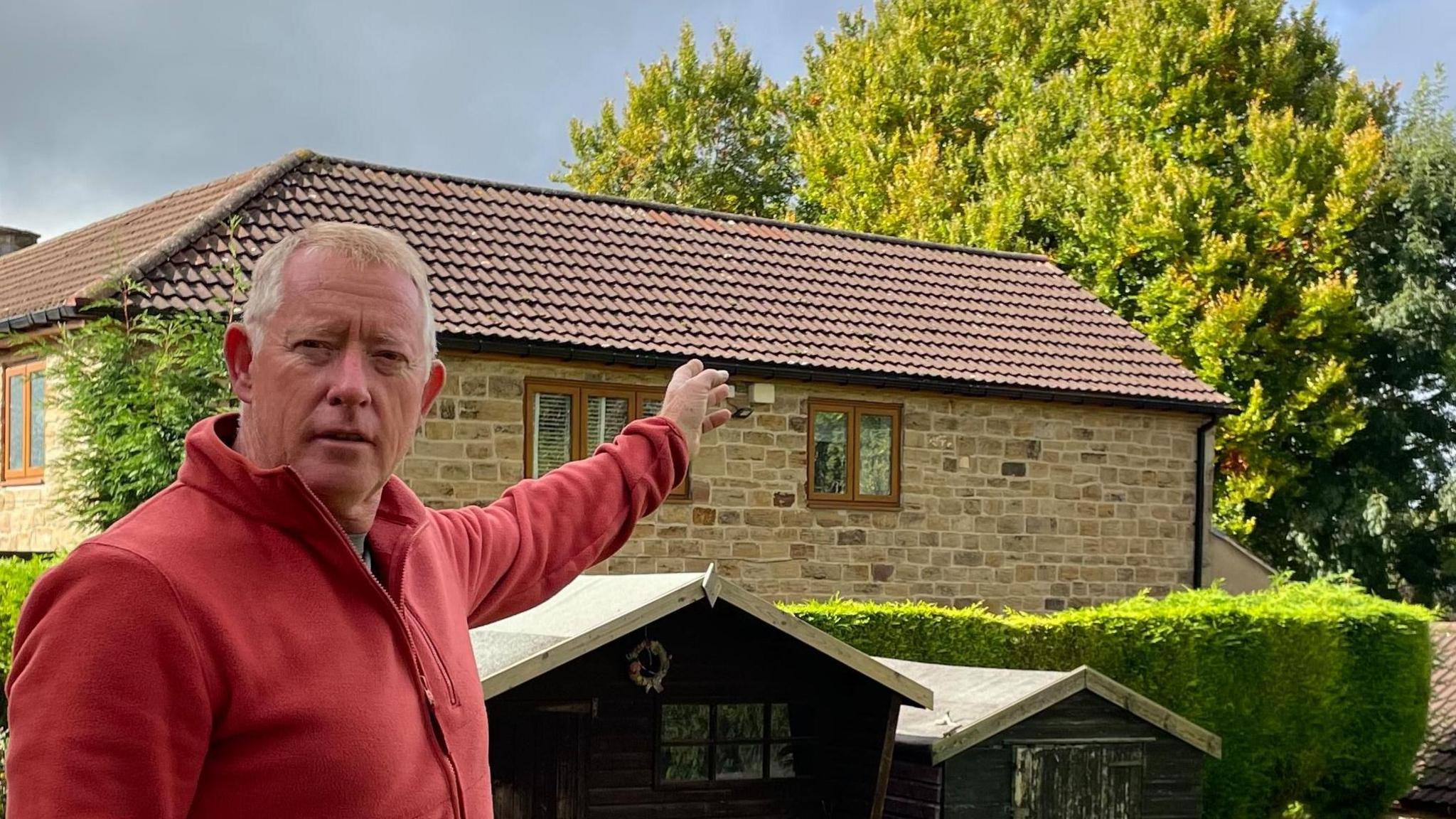 Dave Delany is stood outdoors in his garden. He's pointing towards the beech tree, which is visible behind his neighbour's house.