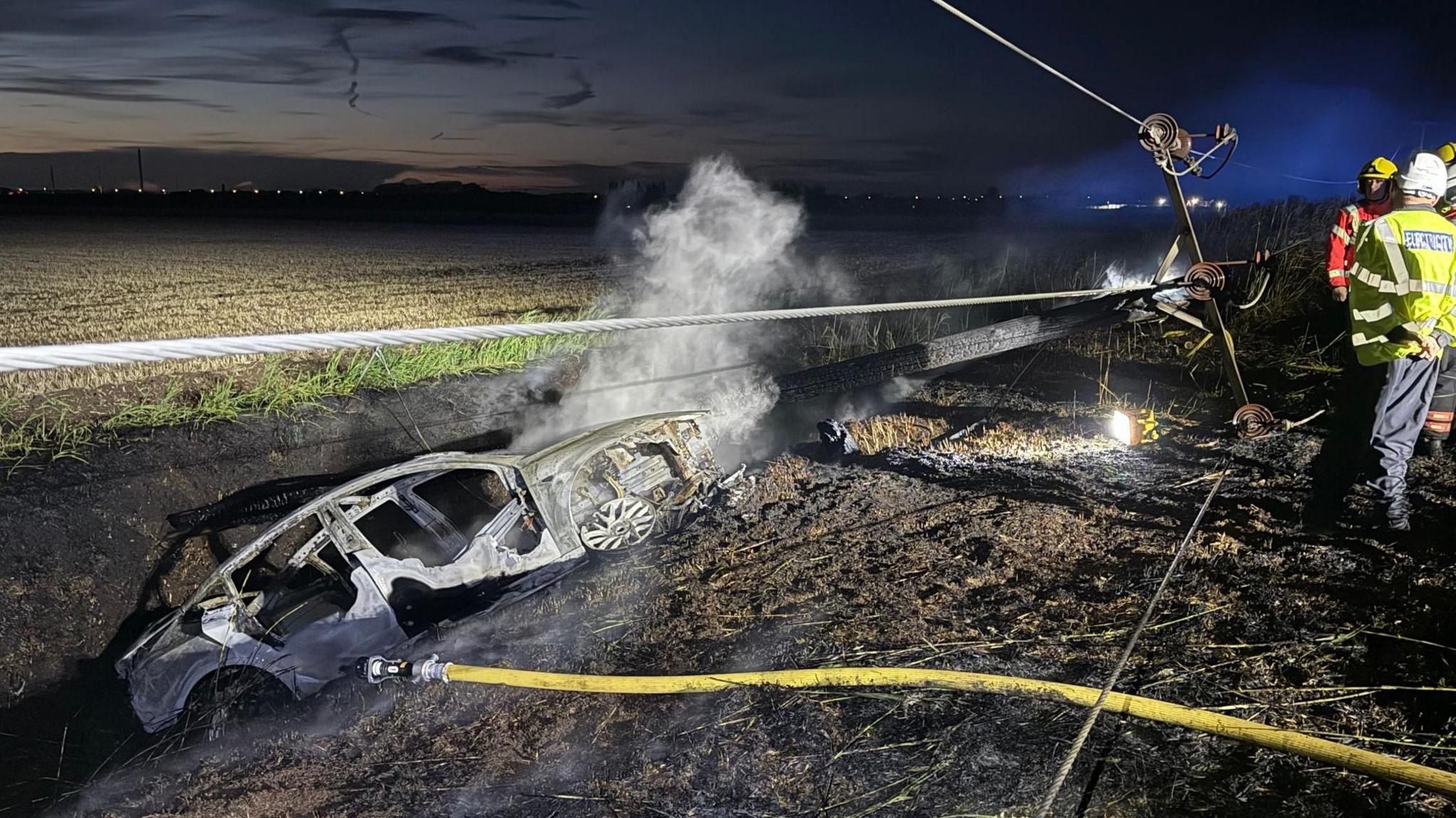 A burnt out car in a ditch next to a field. It is lit up by flood nights in a night time image.