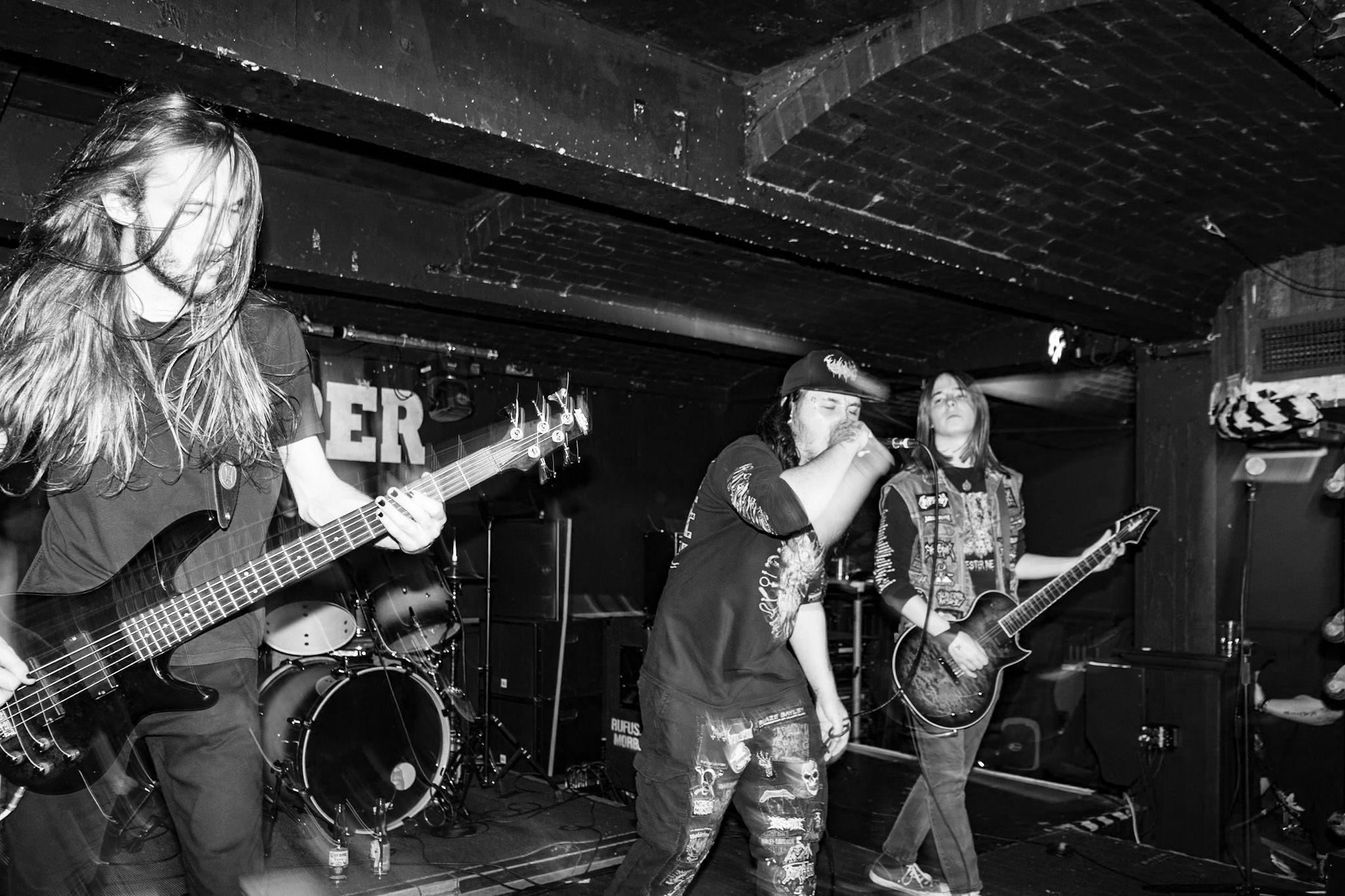 Three members of Morbid Atrocity are seen in a black and white image. The singer, wearing a dark cap, tee-shirt and trousers, is flanked byy two guitarists, both of whom have long hair.