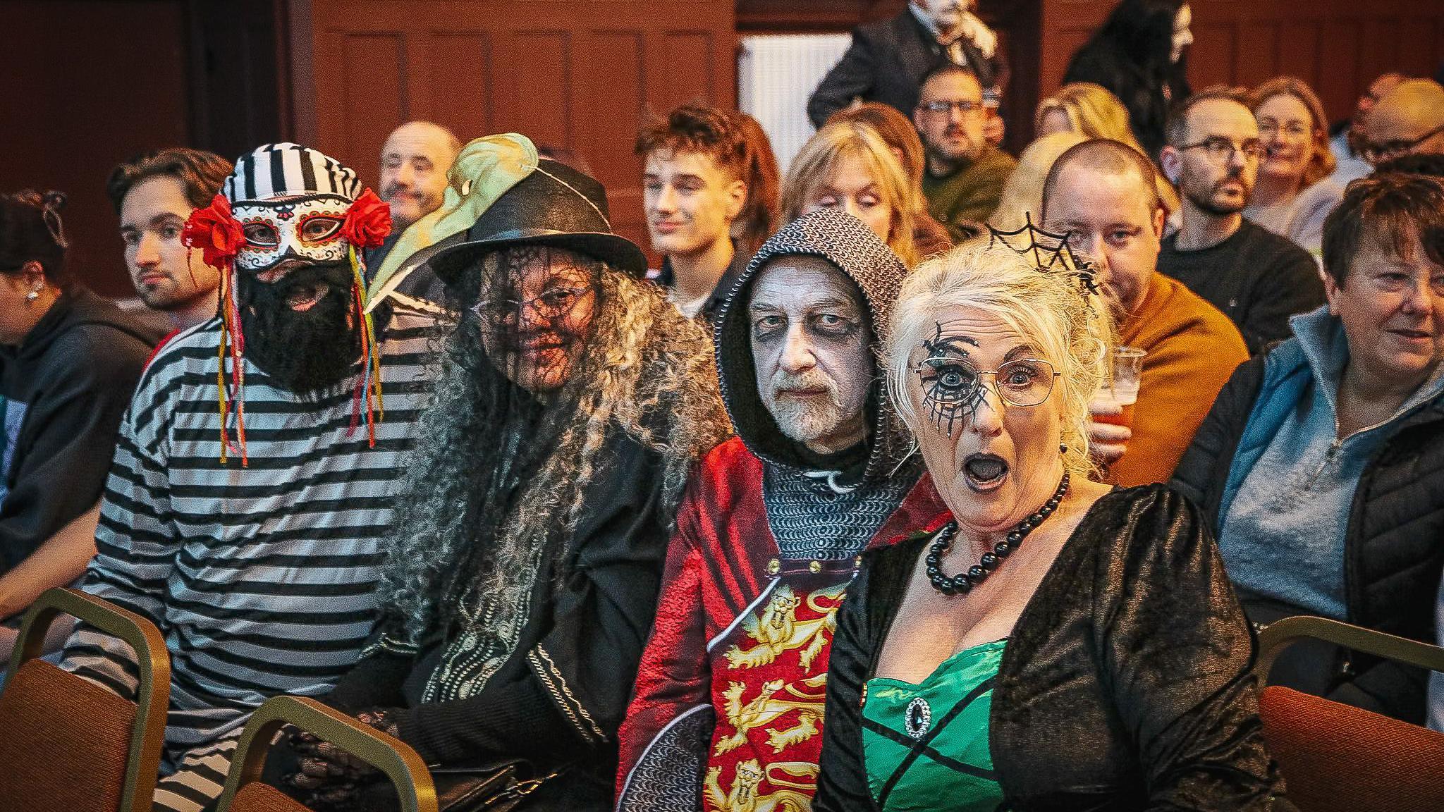A group of people are sat in an row as part of an audience in full Halloween costumes including a witch and a knight. They are posing for the camera.