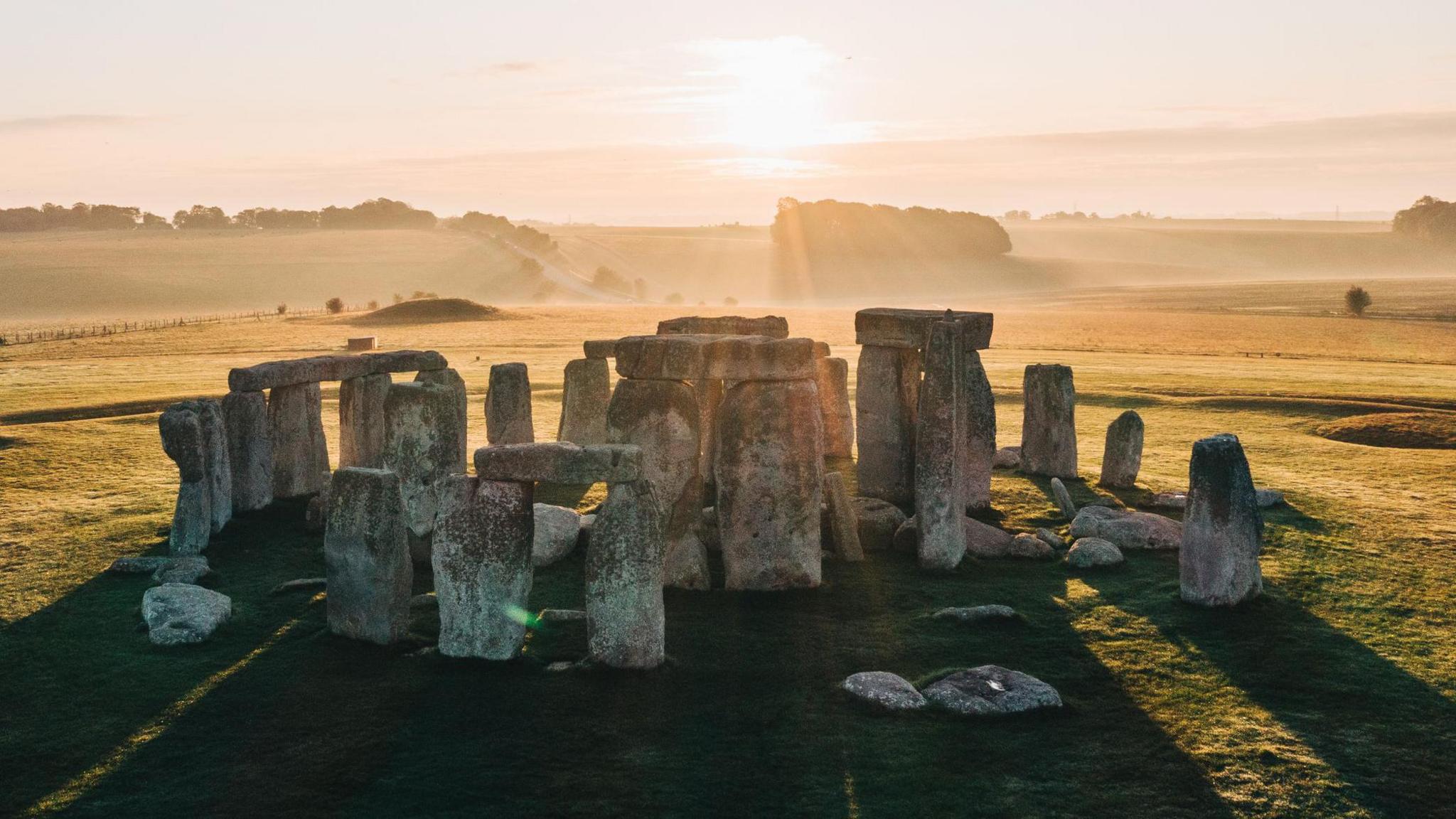 Stonehenge seen with the sun behind and sun beams streaming through the large stones placed in a circle and lighting up the hills and fields behind.