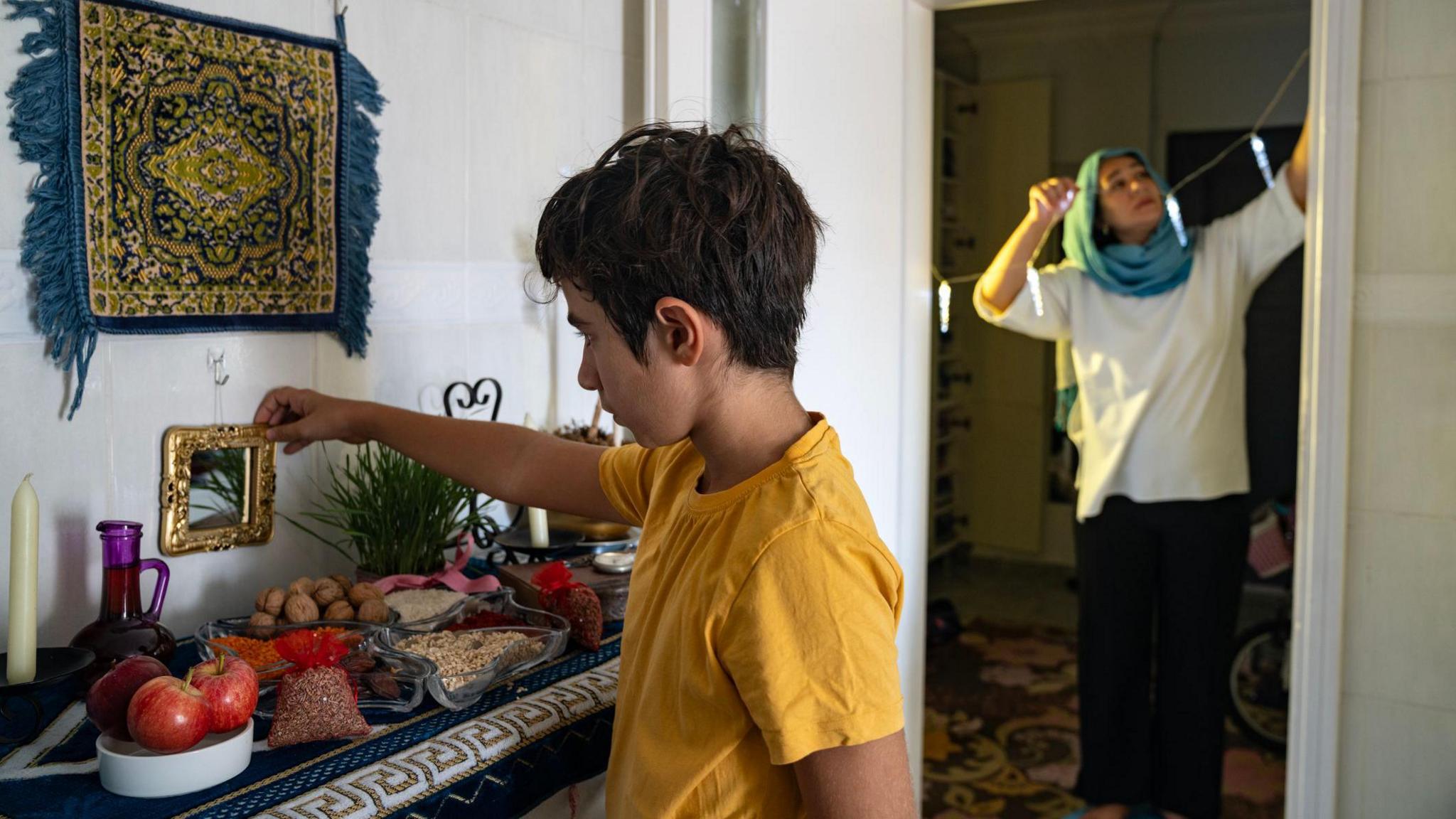 A boy and his Mother at home. In the foreground the boy is adjusting a mantelpiece display of apples, nuts and decorative items. In the background his Mum hangs a garland in a doorframe.