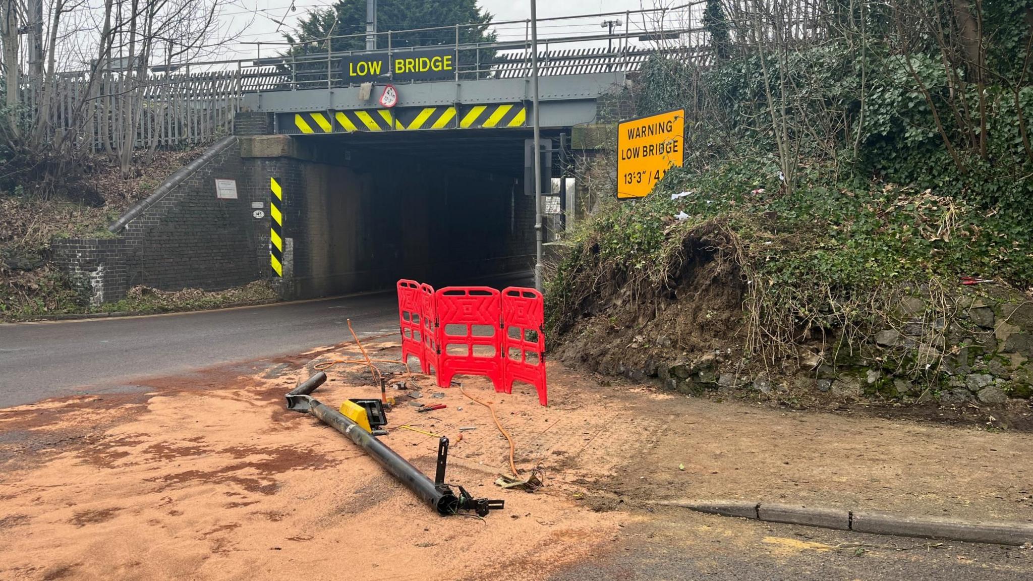 Sand covers the road and pavement. A pedestrian crossing pole lies on its side in front of red plastic safety barriers and low wall on a railway embankments which rises up to the tracks. A road bridge under the railway is in the background. The road under the bridge appears clean, with the sand covering the start of Station Road in Luton, near Leagrave railway station.