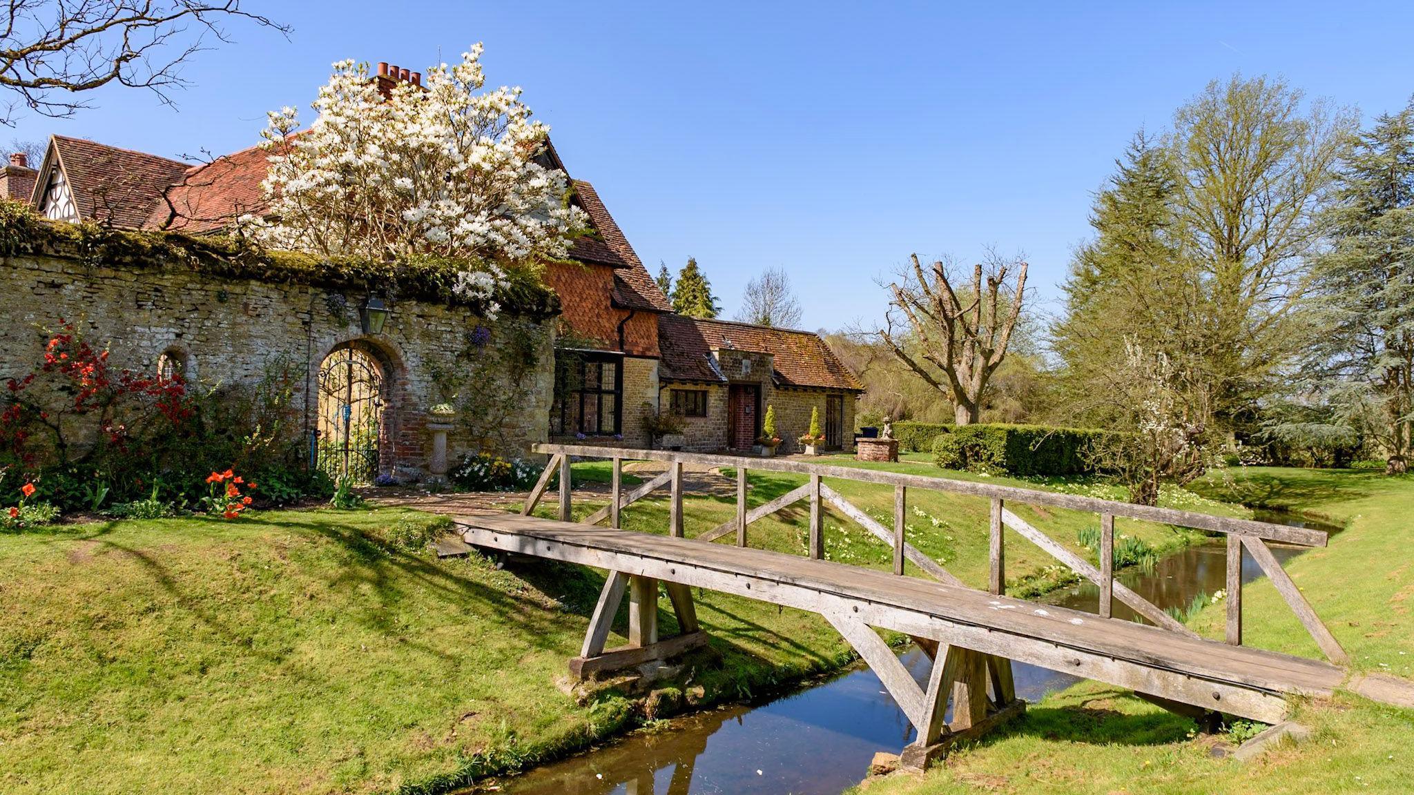Great Tangley Manor, which is an old, stone house with a pointed, red-tile roof. The property is surrounded by manicured grass and other plants, whole a narrow wooden bridge crosses a moat around the perimeter.