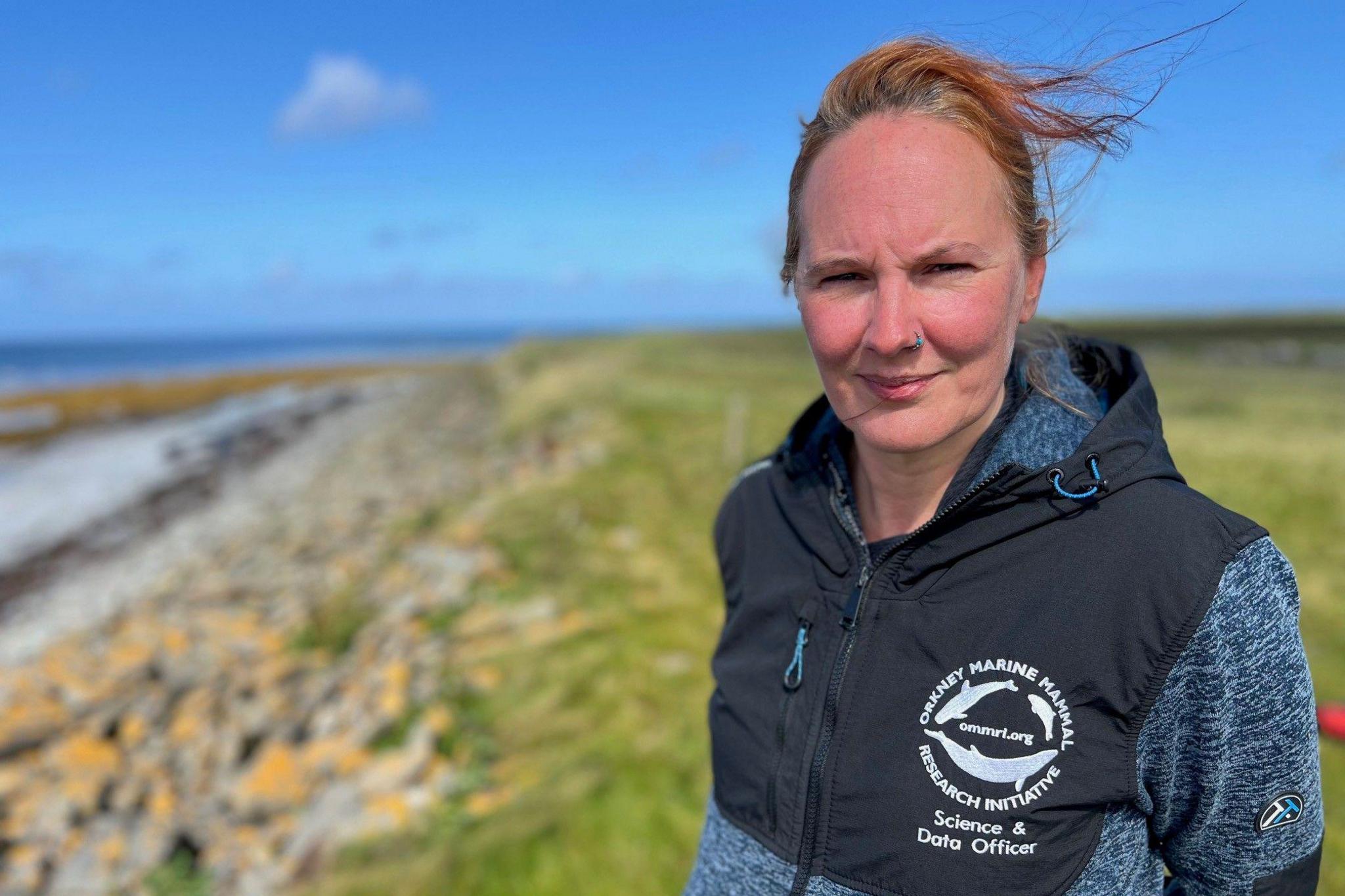 Emma Neave-Webb, with Orkney Marine Mammal Research Institute science and data officer top on, looking at camera at a beach