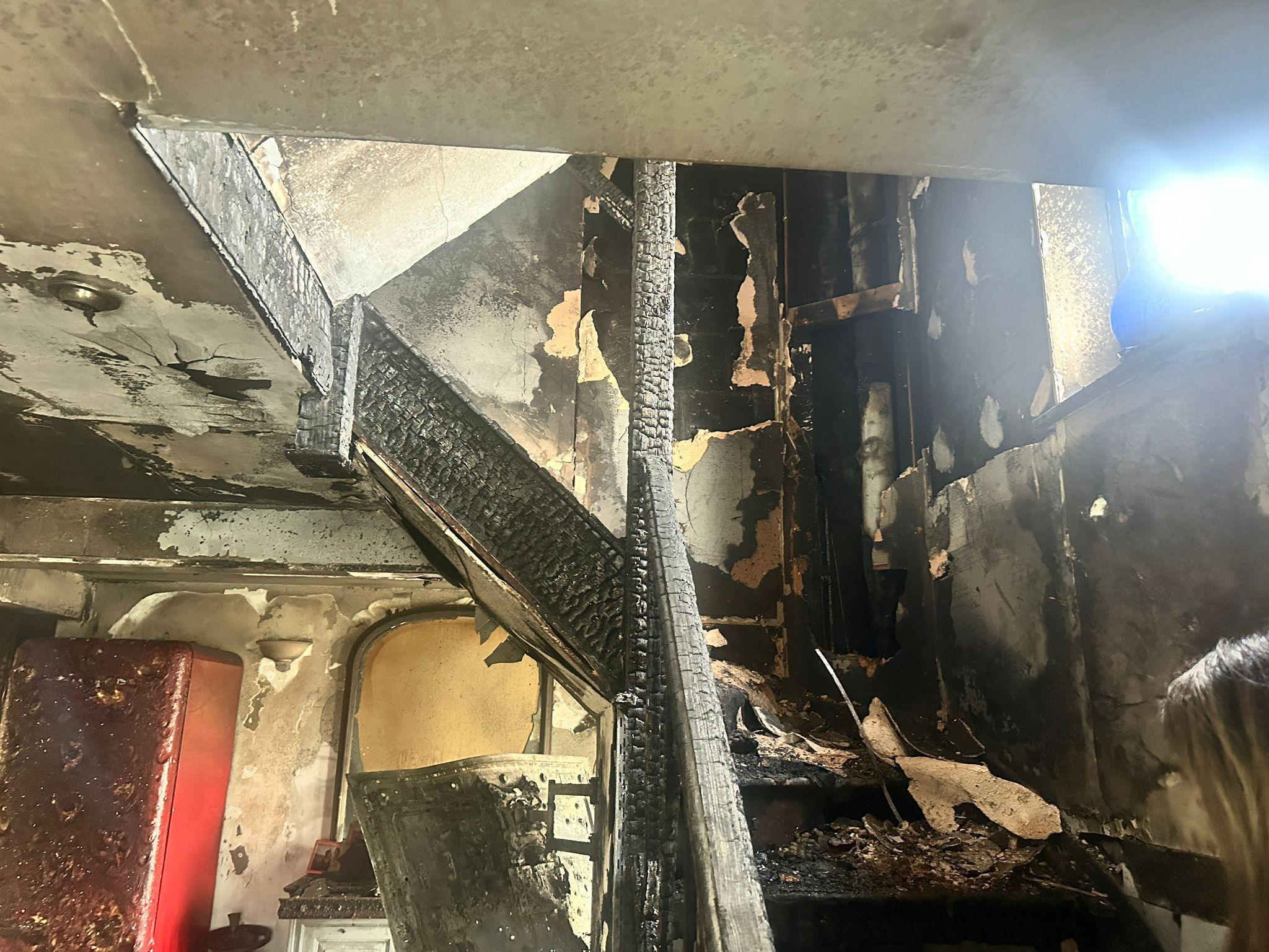 The staircase of the house. It is blackened and burnt. The charred staircase, landing and hallway can be seen. There is wallpaper falling off and the ceiling is cracking from the smoke and fire damage. 