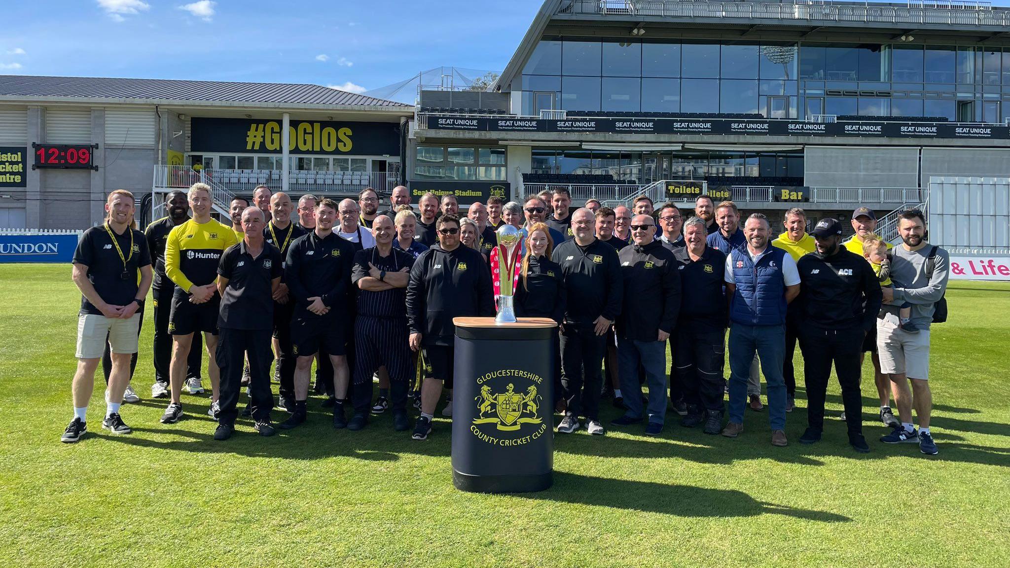 Staff and players of Gloucestershire County Cricket Club stand for a team shot on the pitch at the County Ground in Bristol behind a plinth on which rests the Vitality Blast T20 trophy which they won at finals day at Edgbaston in Birmingham. It is a sunny day and the pavilion is visible in the background.