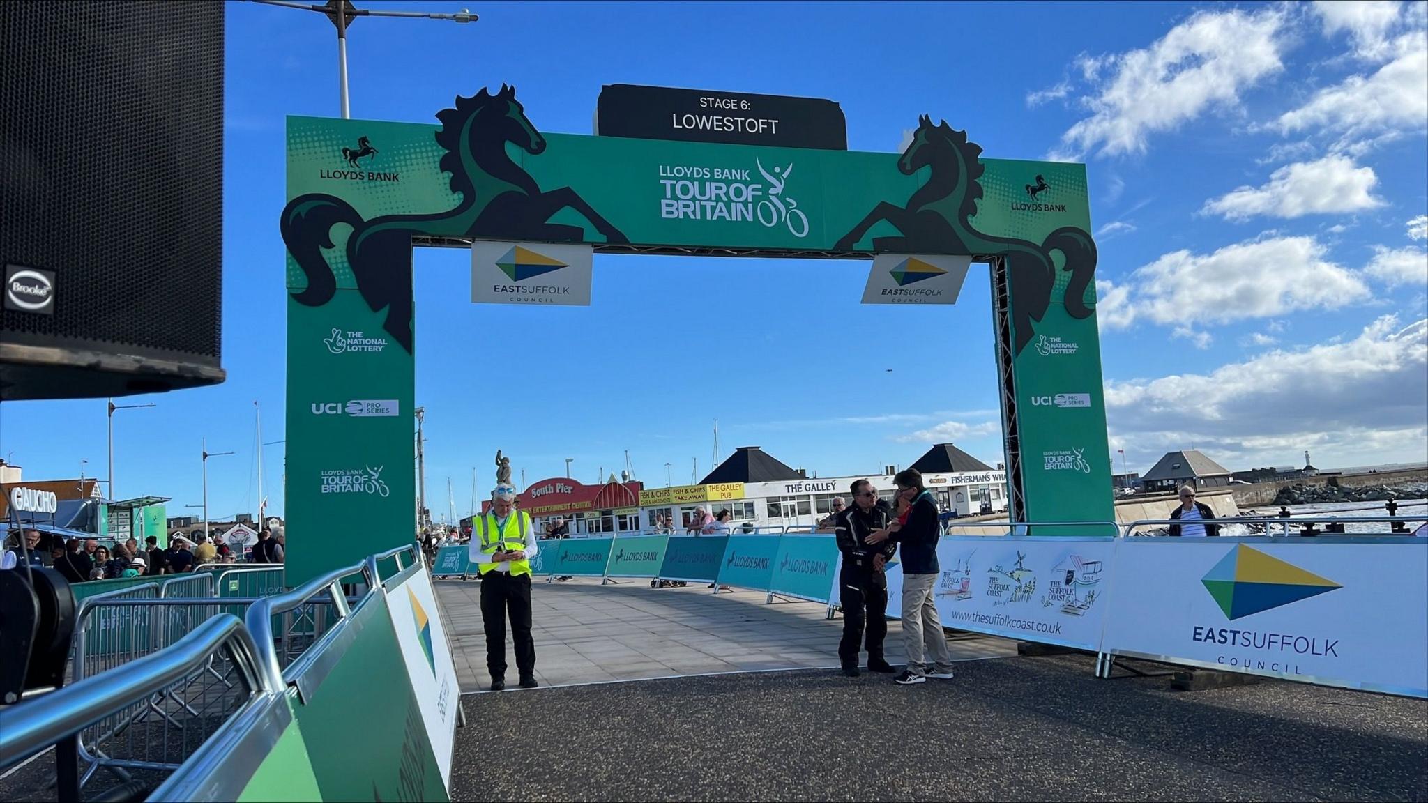 A green Lloyds Bank Tour of Britain banner over a paved surface with Lowestoft's South Pier visible behind. There is a blue sky with white clouds and several spectators and race officials standing around.