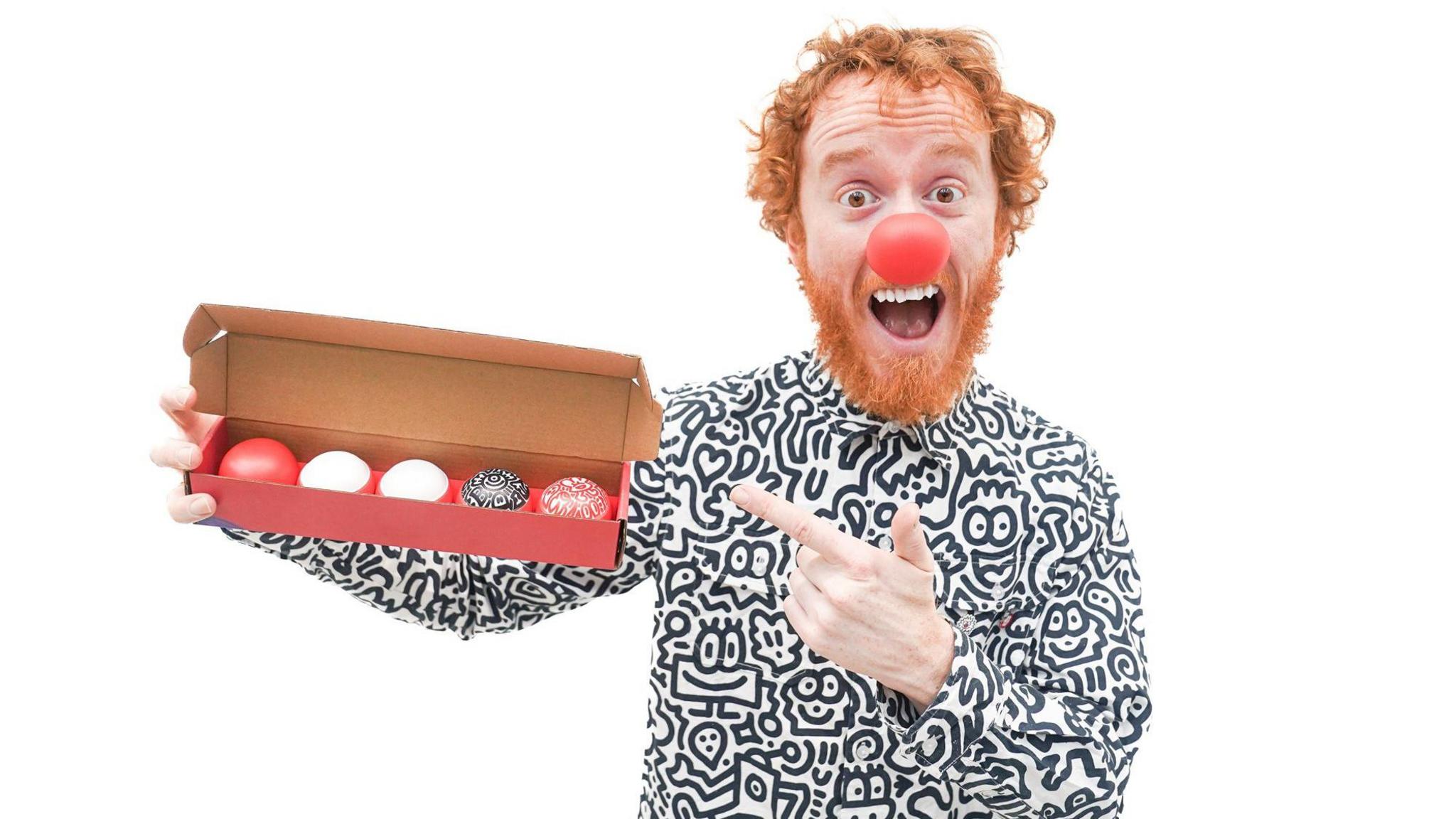 A red-haired man wearing a black and white doodle-designed shirt. He is holding a box containing five noses different designs. He is also wearing a big red nose.