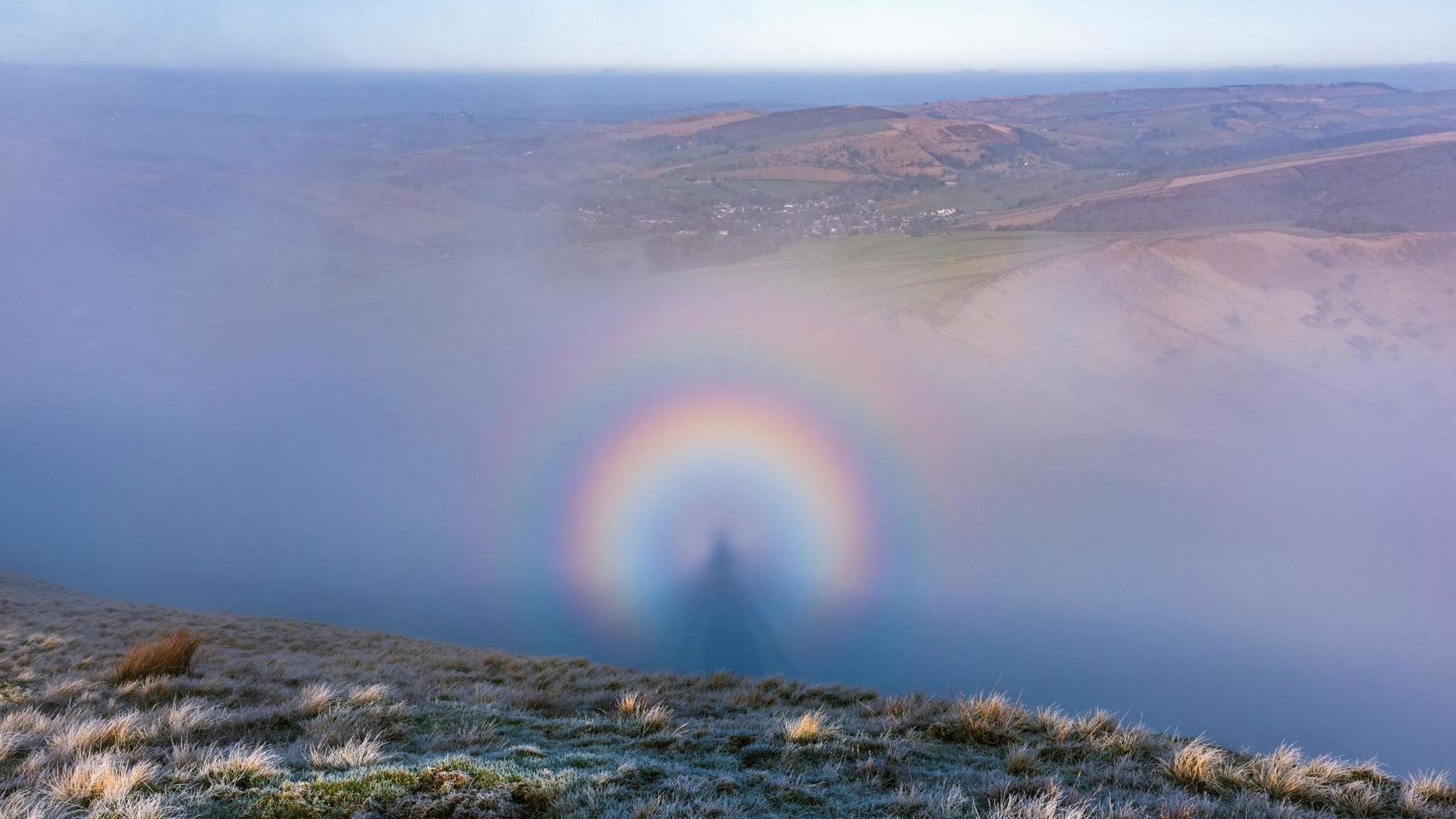 A Brocken spectre, with the shadow of a person surrounded by a circular rainbow, can be seen in the mists in a valley off a frosty, grassy ledge