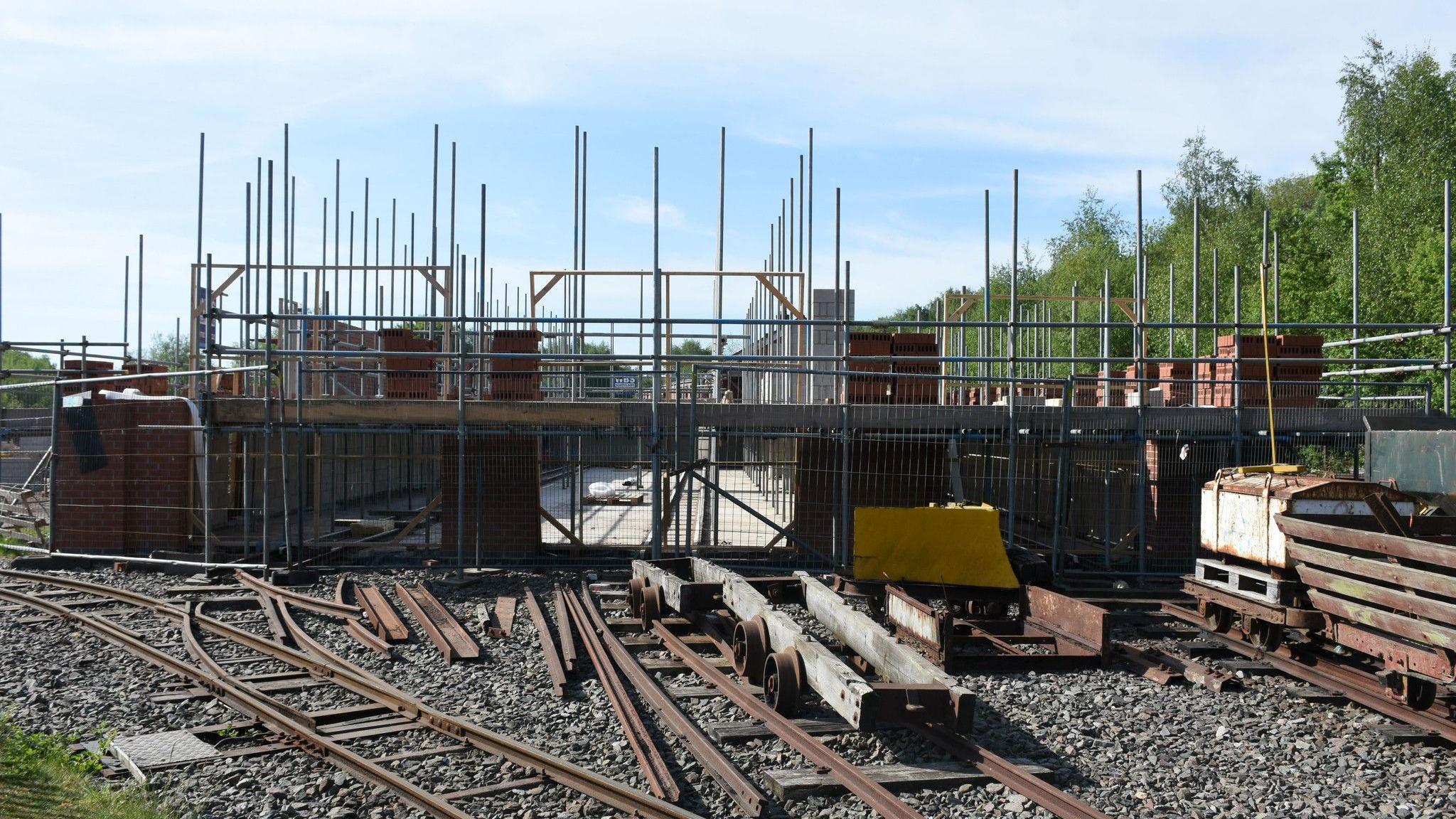 A series of railway tracks lead up to a construction site which comprises scaffolding and a brick-built structure. There are various pieces of railway equipment on and alongside the tracks.