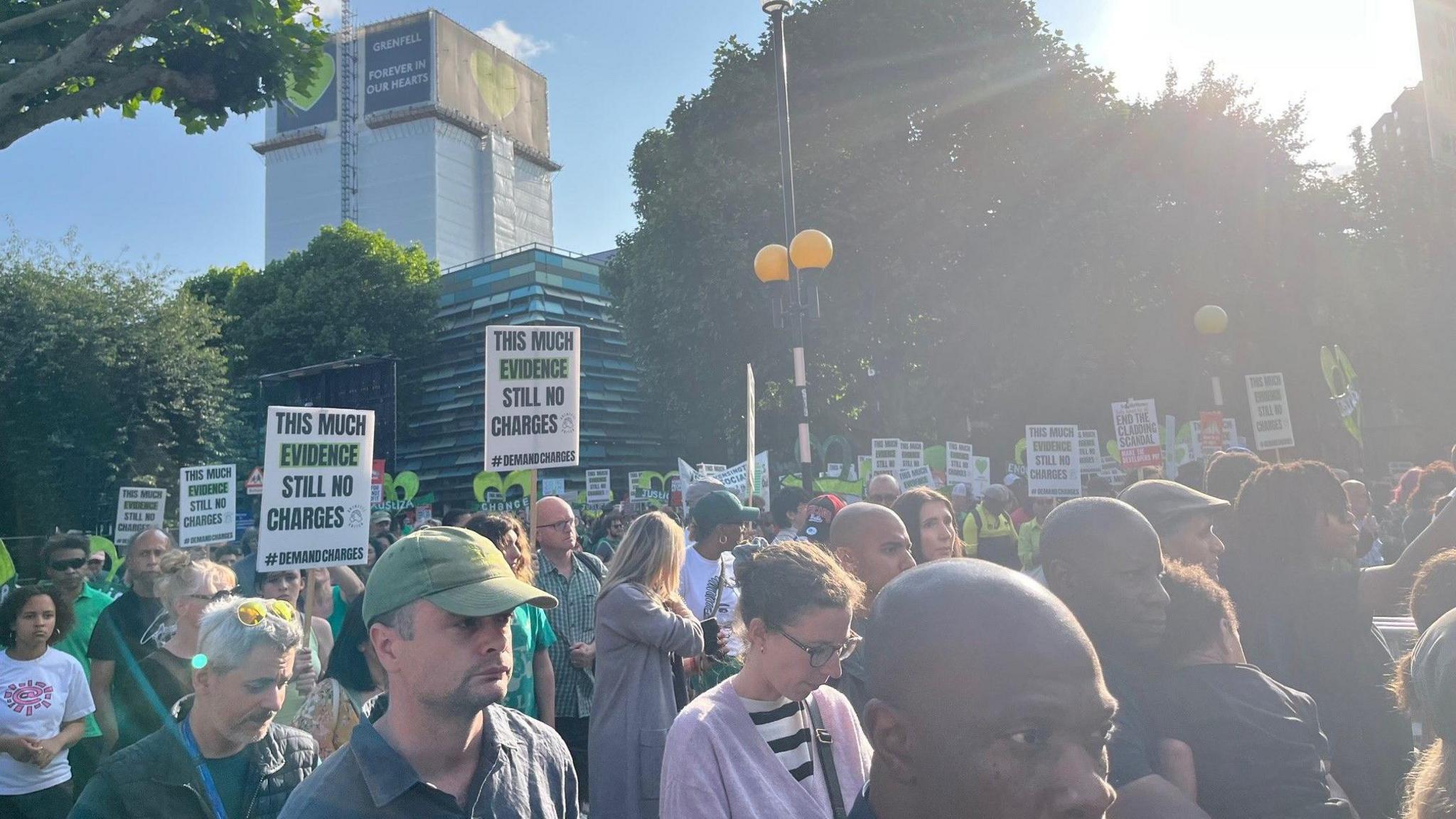 Crowds walking with placards saying 'this much evidence and still no charges'. In the background is Grenfell Tower
