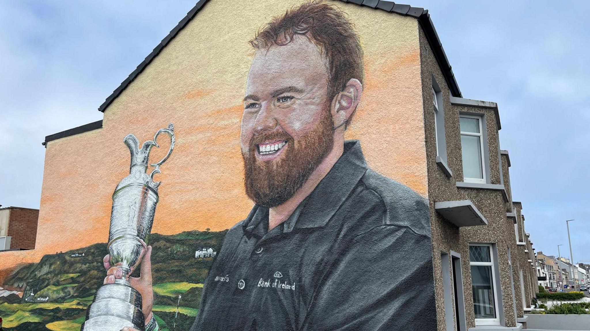 A wall mural showing Shane Lowry wearing a dark Bank of Ireland polo shirt and smiling while holding a silver trophy