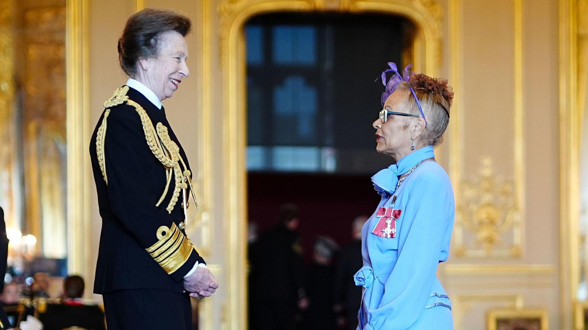 In a room at Windsor Castle, the Princess Royal, who is wearing Royal Navy ceremonial uniform, smiles at Anita Neil, who is facing her. 