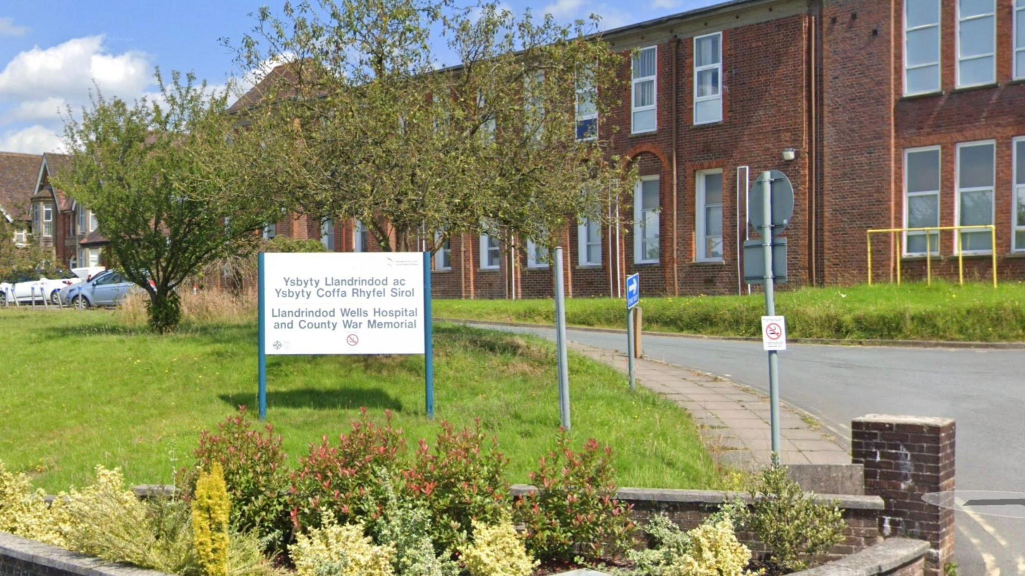 The outside of a red brick hospital building which is long with multiple windows. In front are some trees, a patch of grass and some bushes. On the grass is a white sign with the words Llandrindod Wells Hospital and County War Memorial.