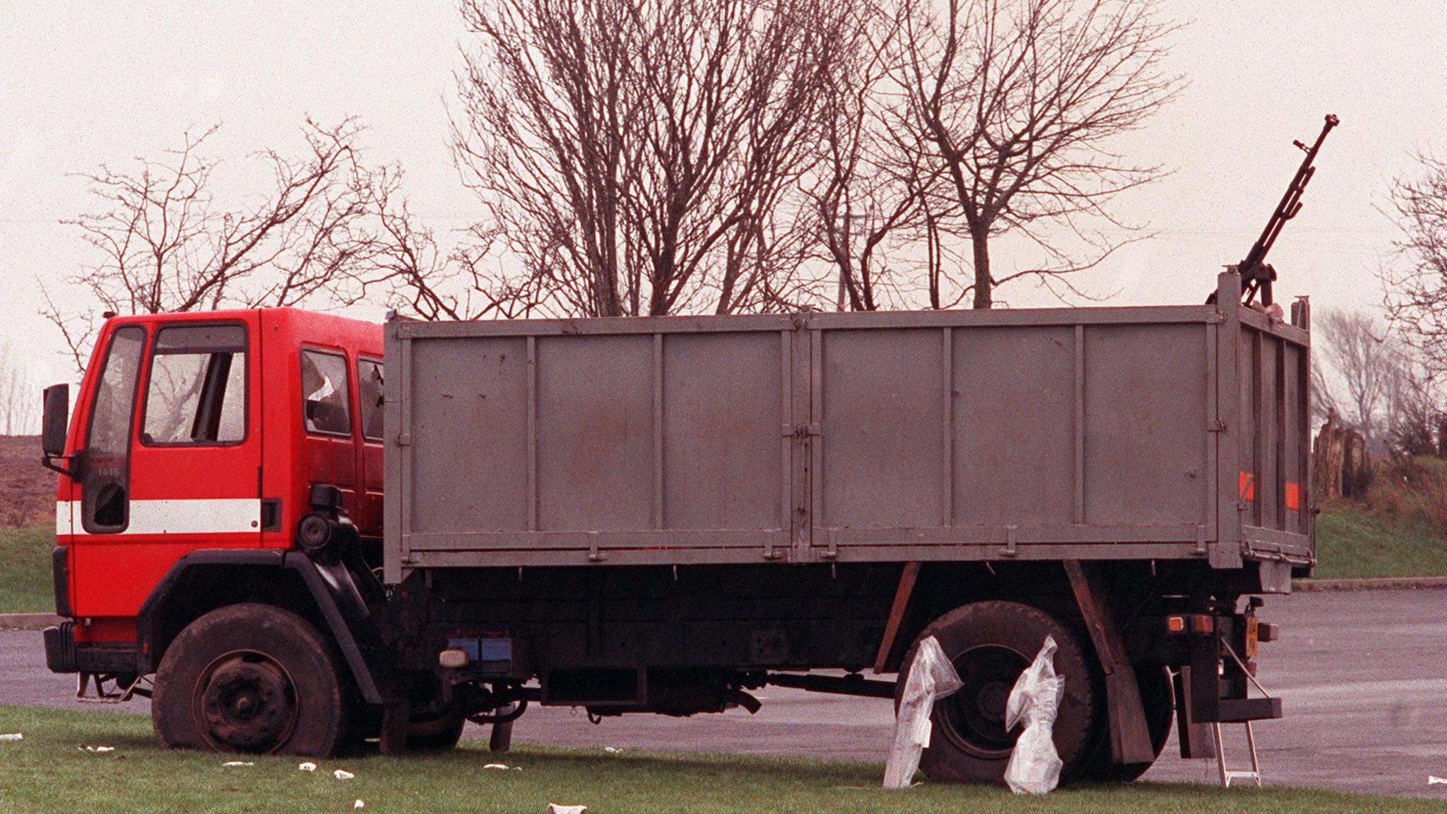 Lorry which had a heavy machine gun welded to its tailgate. It is painted red with a white stripe across the front. The grass before it is littered with white strips and the trees are bare across a grey sky. 