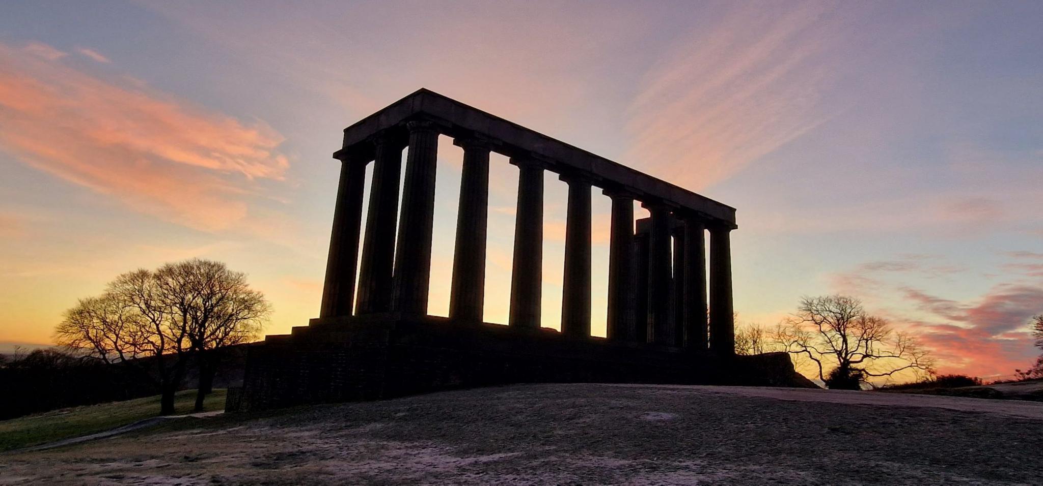 The monument, which comprises a number of stone pillars, is silhouetted against a sky filled with colours of pink and blue.  