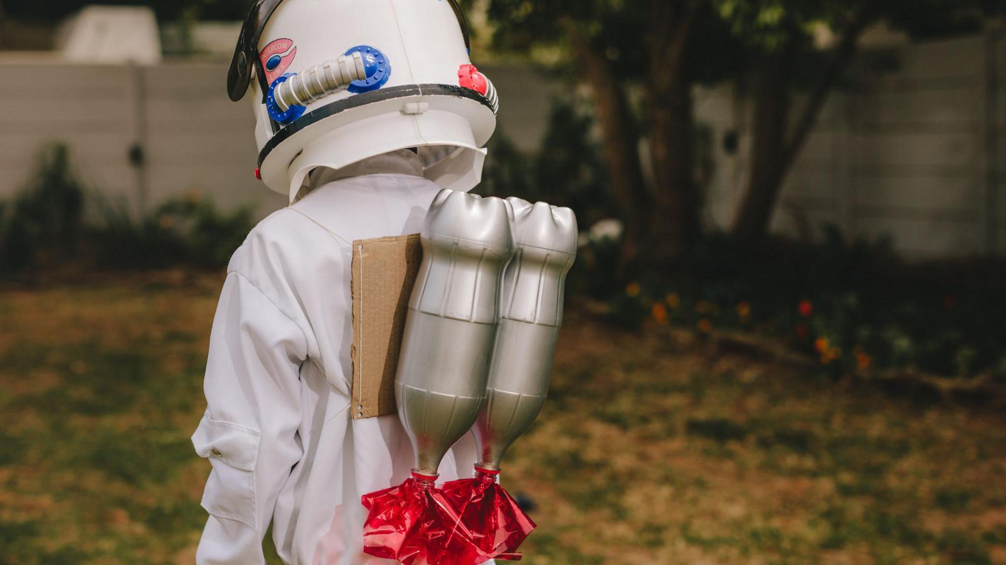 A young child with their back to the camera wearing an astronaut costume and a rocket pack made of silver spray-painted plastic bottles