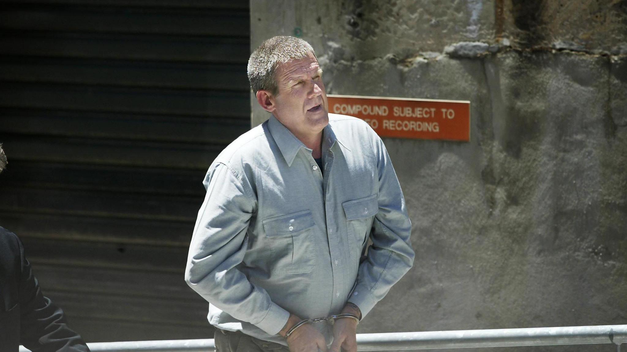A man in his forties walks in handcuffs. Behind him is a large stone wall.