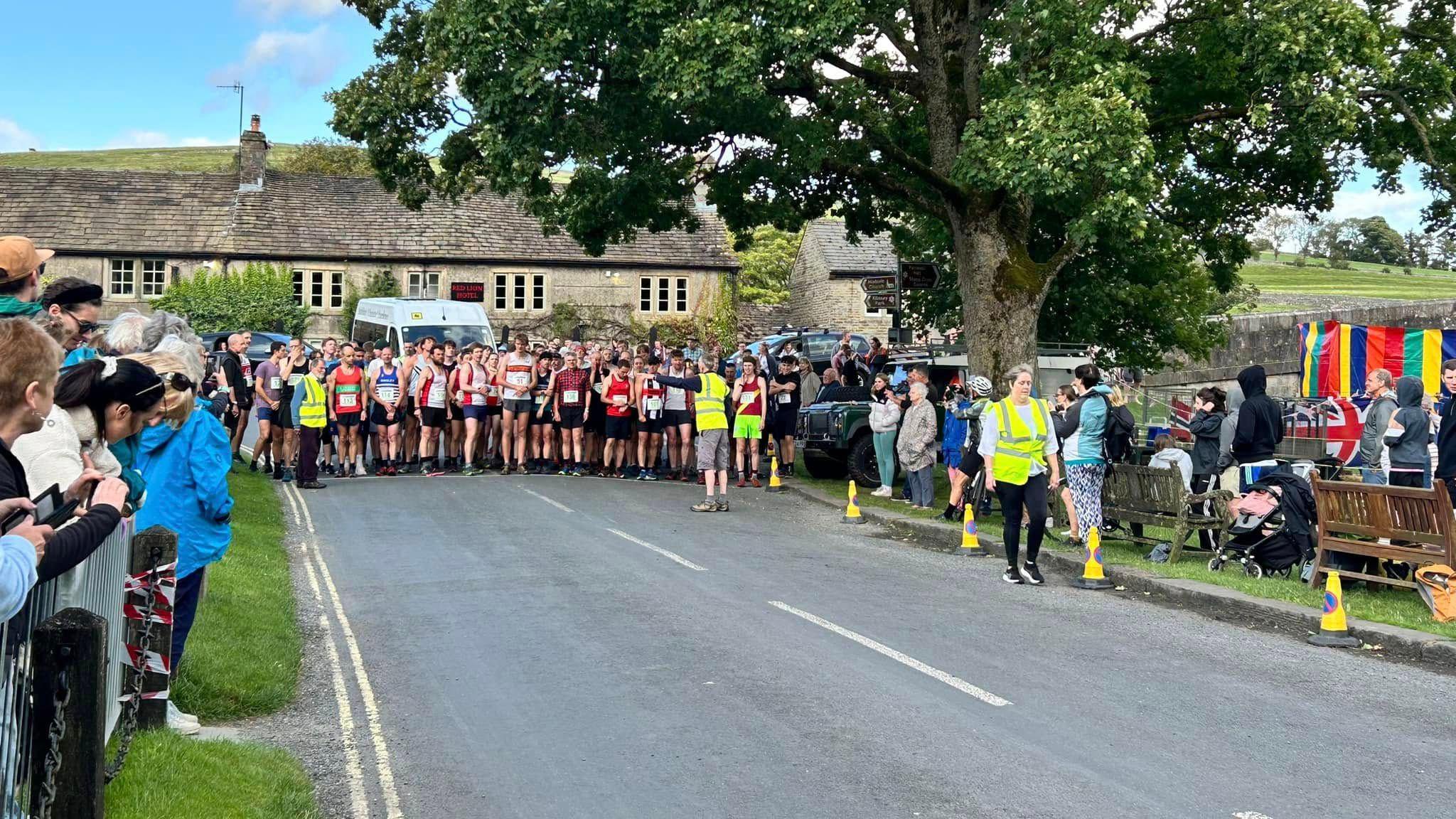 A group of runners amass on a road in front of a country pub. Crowds line the road. There are trees to the right and marshals with yellow vests.